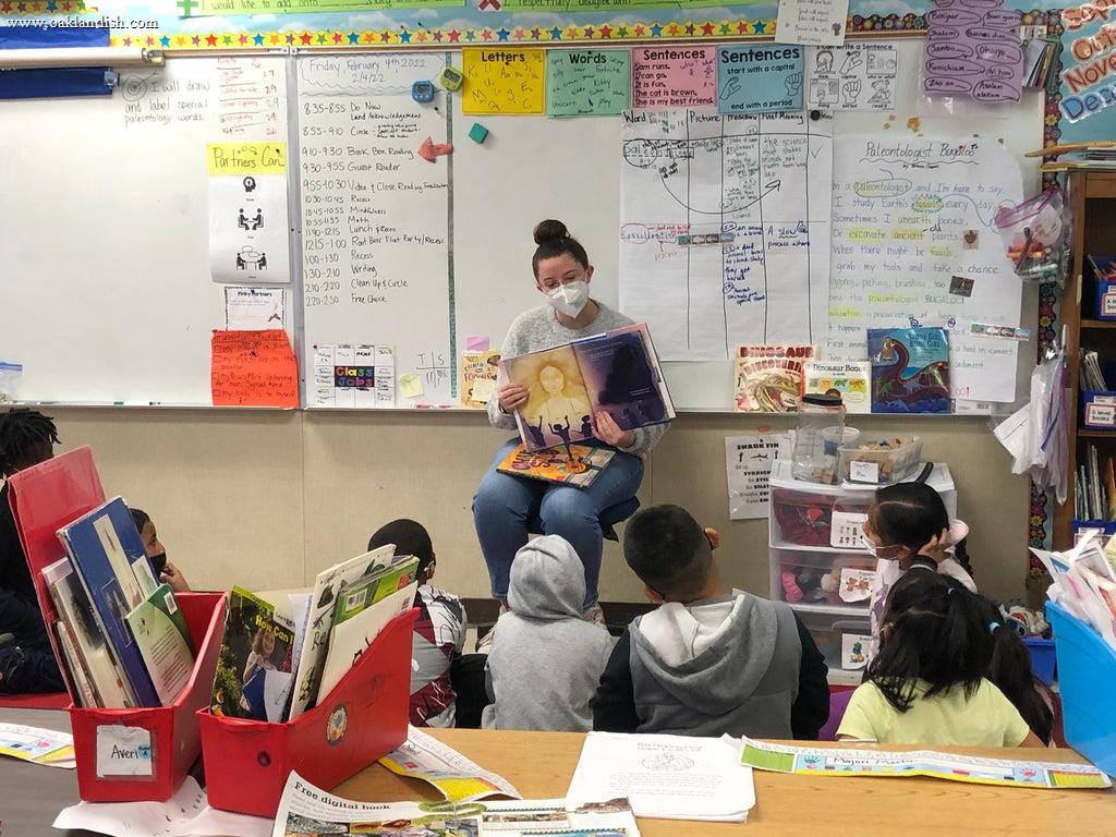 teacher in classroom, seated, reading to kids sitting on floor