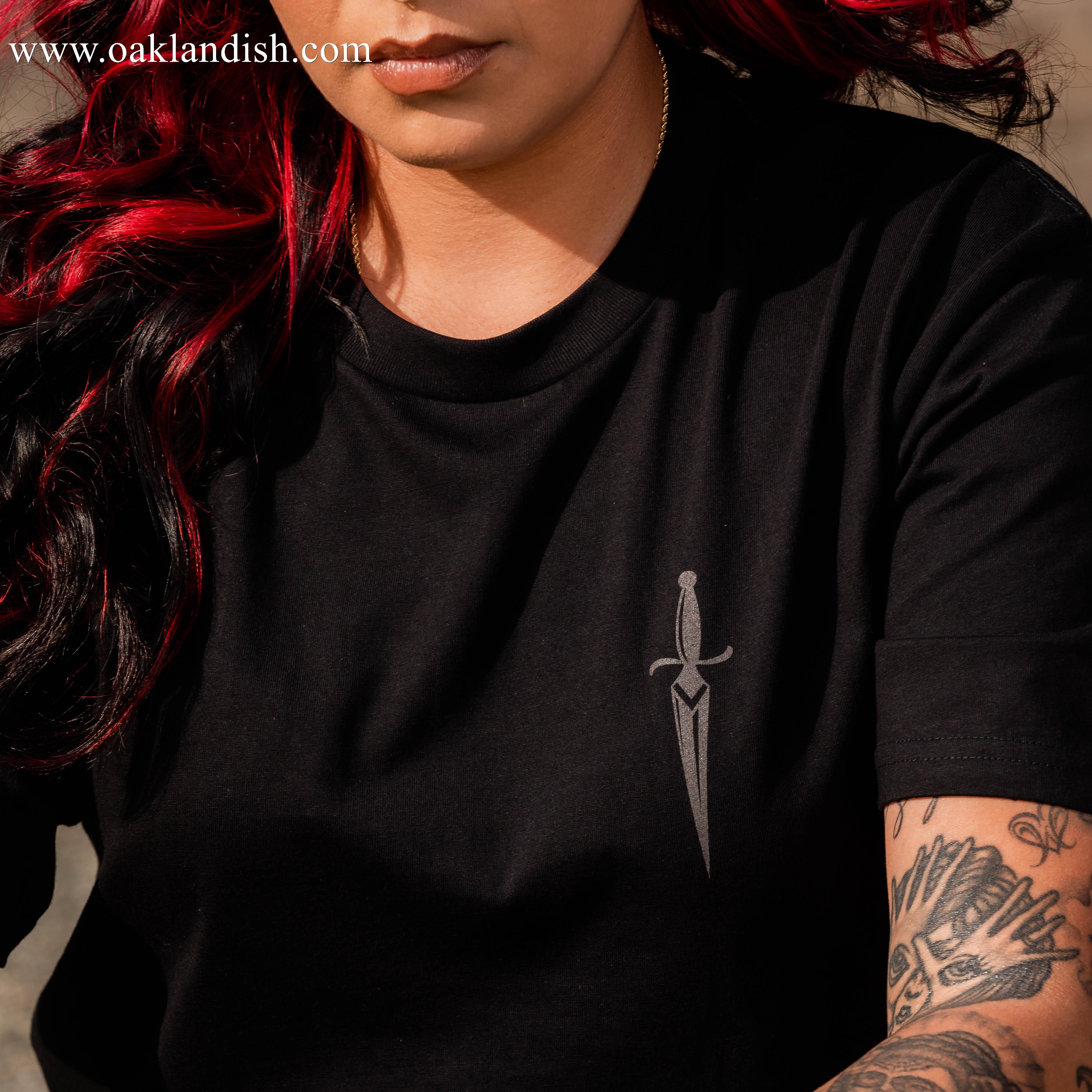 A person with long, dark hair featuring red highlights is wearing the Blossom Tee by Oaklandish—a men's cotton t-shirt adorned with a small sword graphic on the left side of the chest. Their left arm is decorated with various tattoos, including floral motifs and other intricate patterns. The person is sitting outdoors.