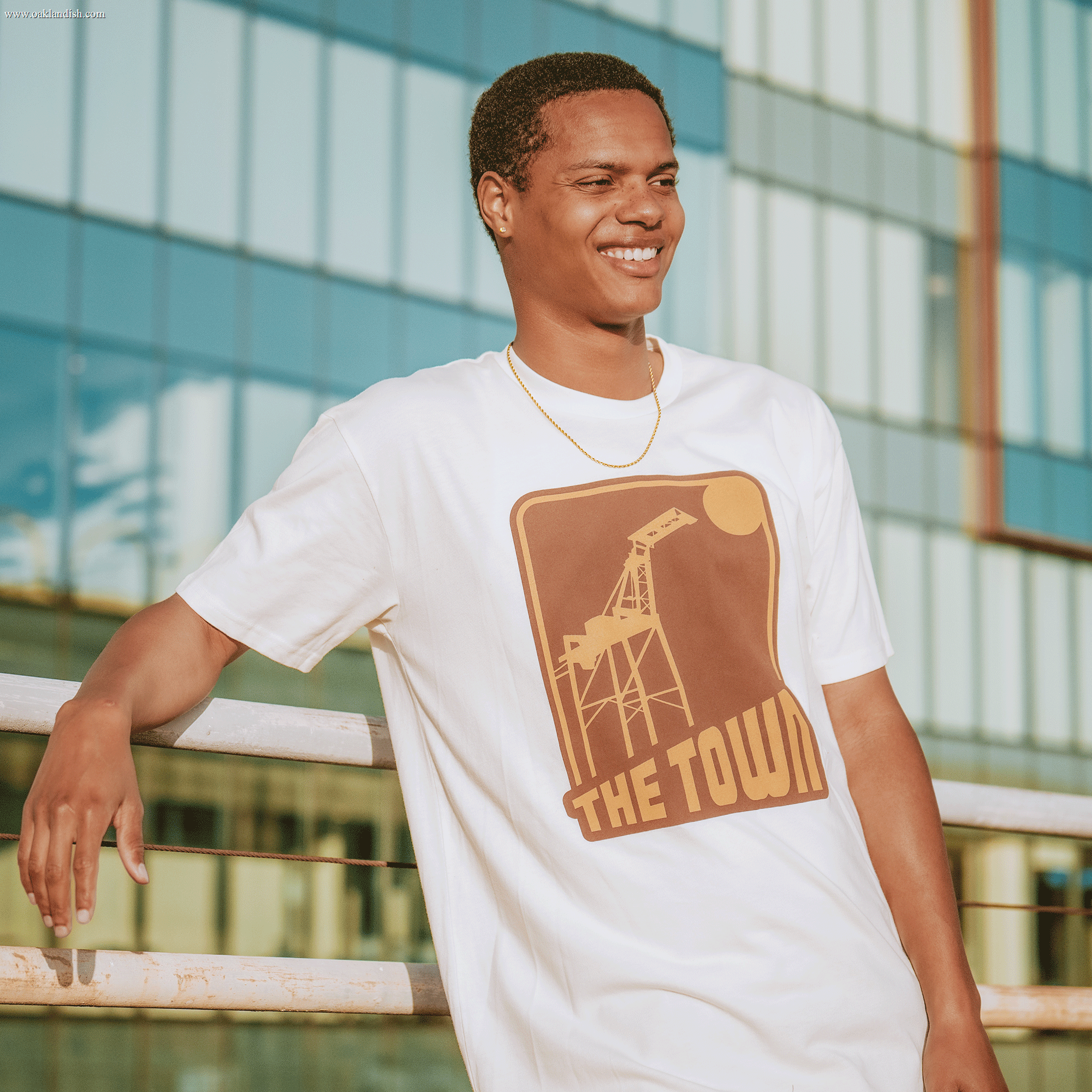 A smiling man leans on a metal railing, wearing the 70's Trading Card Tee by Other—a white t-shirt featuring a vintage-inspired brown graphic of an oil pump and the words "THE TOWN." With his short hair and gold chain, he stands in front of a modern glass building in sunlight. This look effortlessly blends everyday style with retro streetwear vibes, making it perfect for any fan with an appreciation for fresh classics.