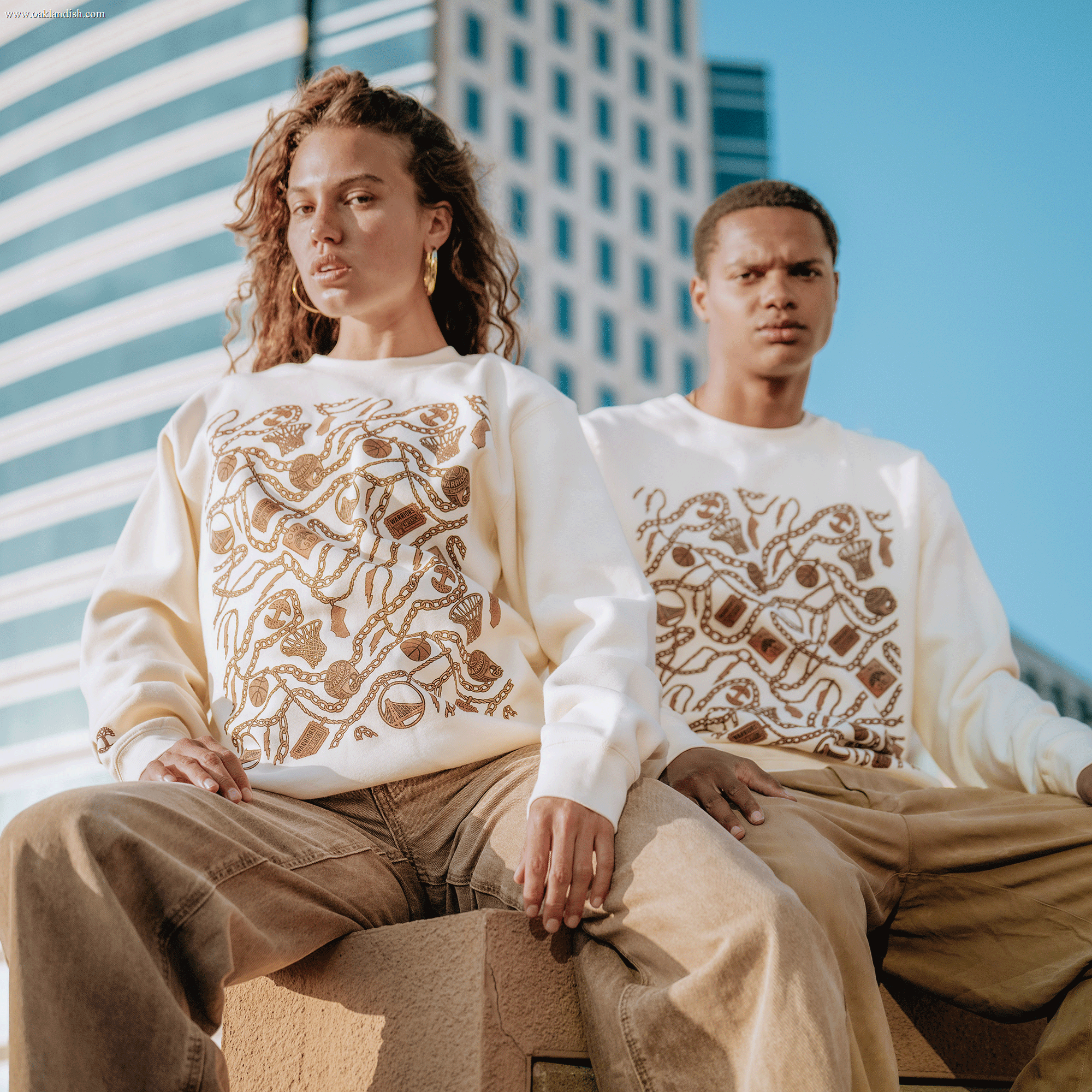 Two people wear cream All Gold Forever Crew sweatshirts by Other, featuring gold snake designs, paired with loose khaki pants. They sit on a ledge outside with modern skyscrapers in the background. Both have calm expressions and sunlight highlights their faces.