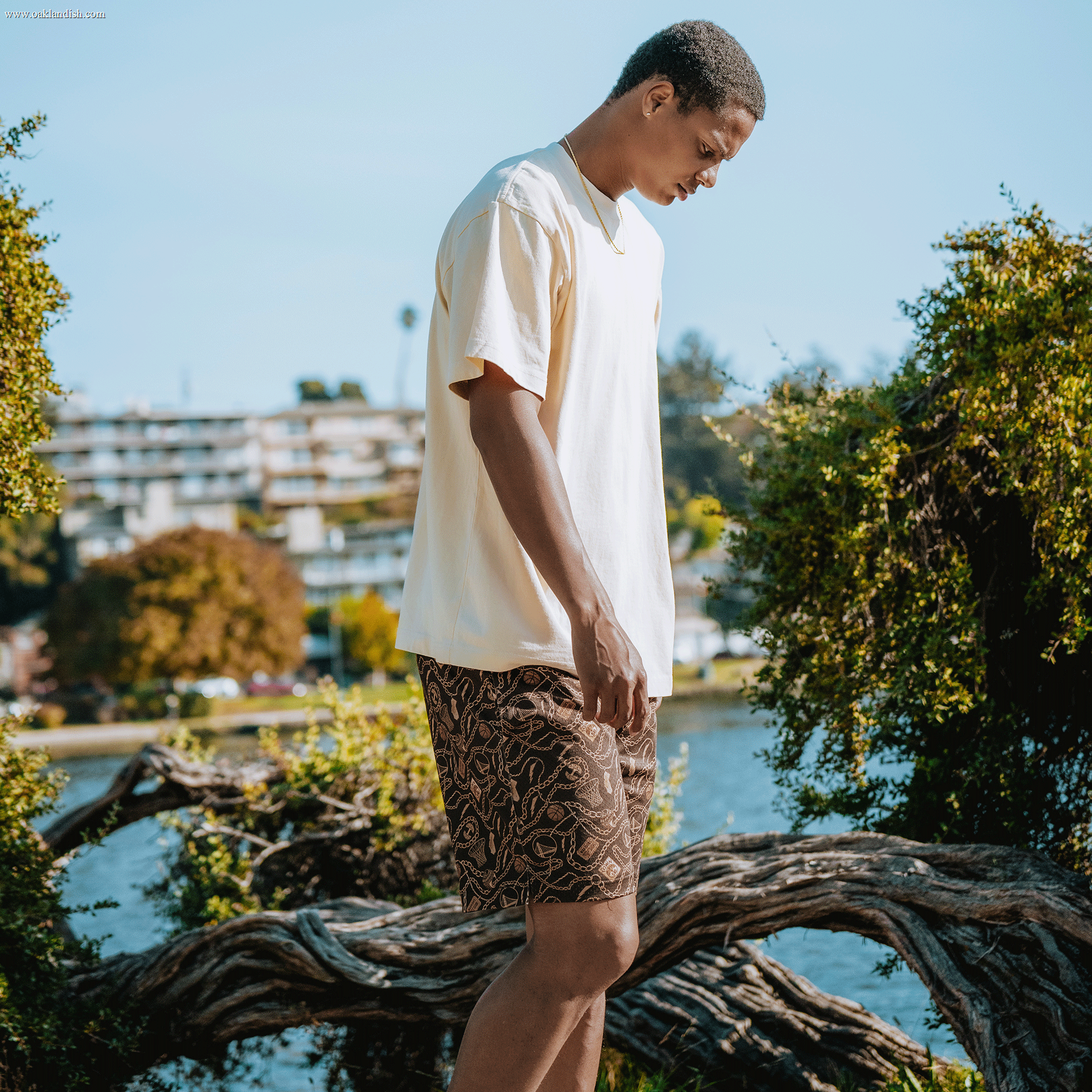 A young man wearing a cream Town Collection t-shirt and All Gold Forever Shorts by Other stands outdoors near twisted tree branches and water, looking down. Bushes and trees surround him, with blurred buildings and a clear sky in the background—a subtle nod to the laid-back spirit of the Golden State Warriors x Oaklandish collaboration.