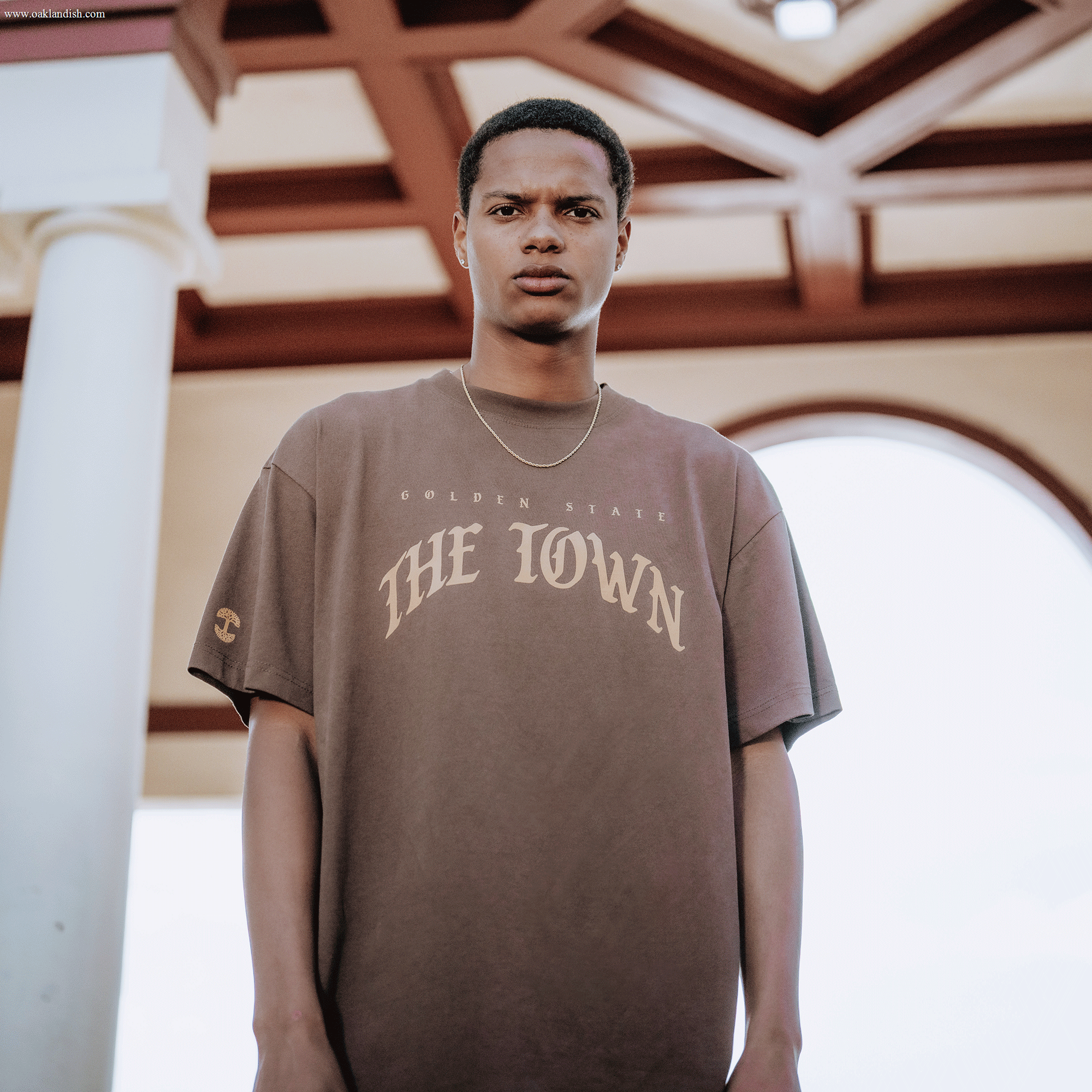 A young man stands outdoors beneath a decorative ceiling, wearing the Other Arched Black-Letter Tee—a loose brown shirt emblazoned with "Golden State The Town"—paired with a thin necklace. He has a serious expression, framed by a white column and an arched window in the background.