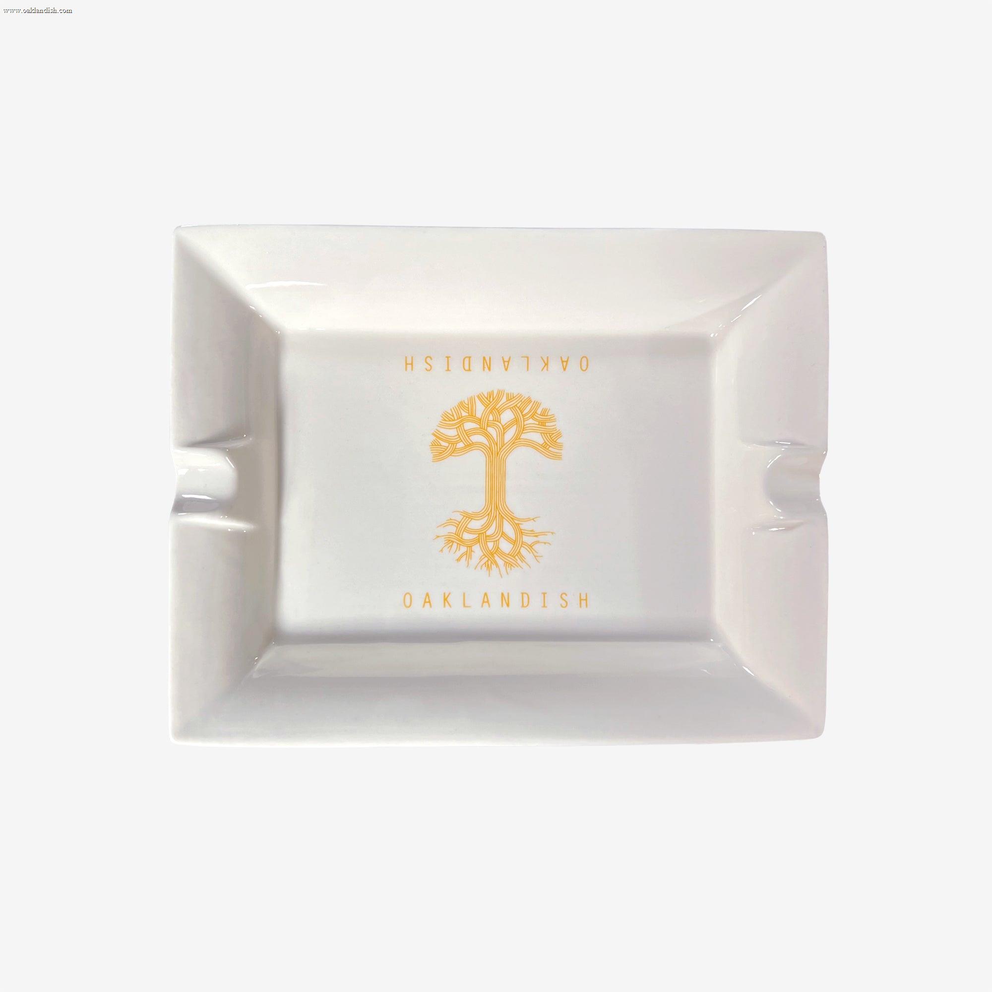 The Oaklandish Ceramic Ashtray by Oaklandish is a rectangular, white ceramic ashtray with a simple design showcasing an orange tree with visible roots in the center. The "OAKLANDISH" logo is imprinted above and below the tree in orange capital letters. Additionally, two grooves on opposite sides of the ashtray are designed to hold cigarettes.