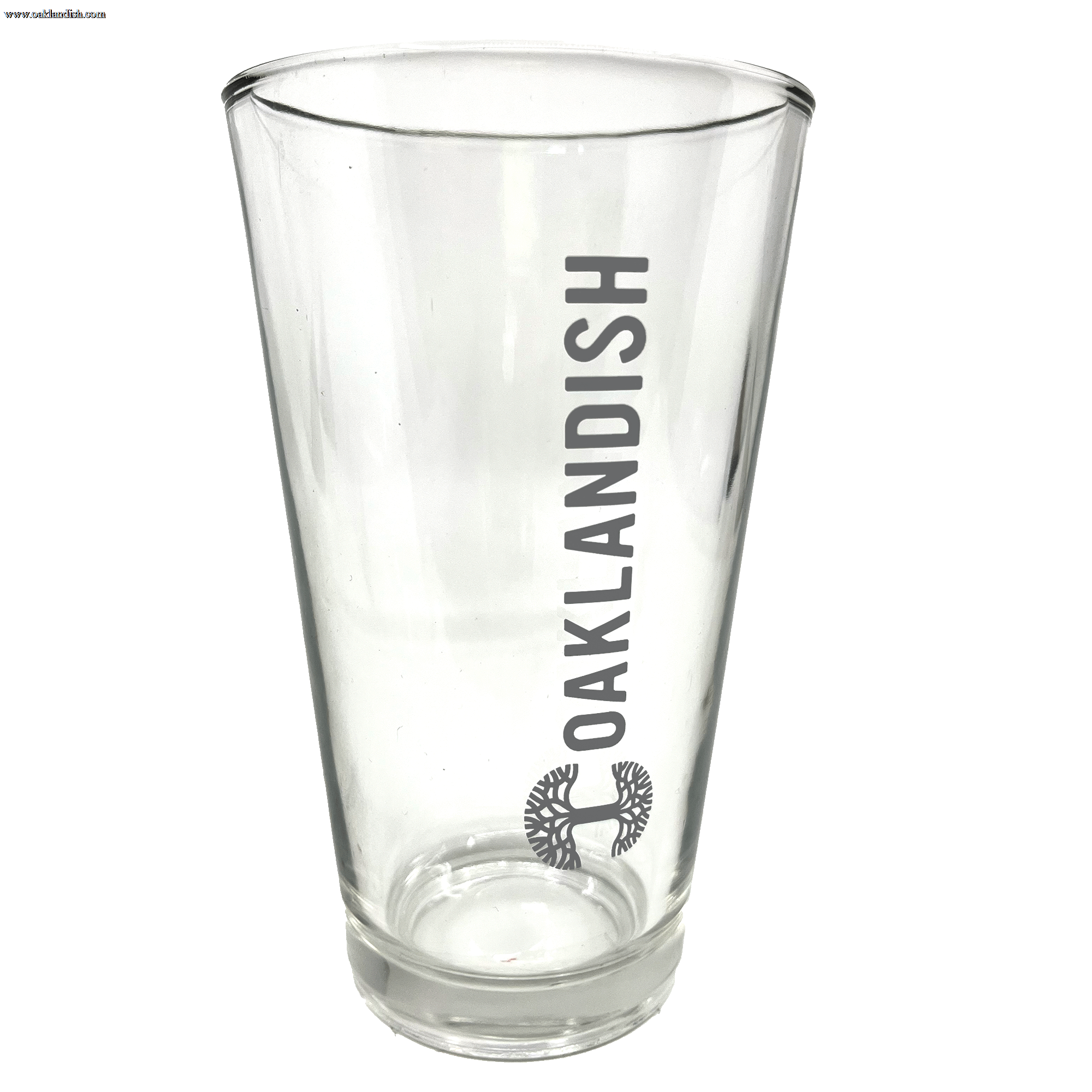 Image of the Oaklandish Banner Pint Glass—a clear, tapered drinkware item featuring the dark grey Oaklandish logo printed vertically. Below the logo is a black image of a spreading oak tree. The background is entirely white, emphasizing the pint glass.