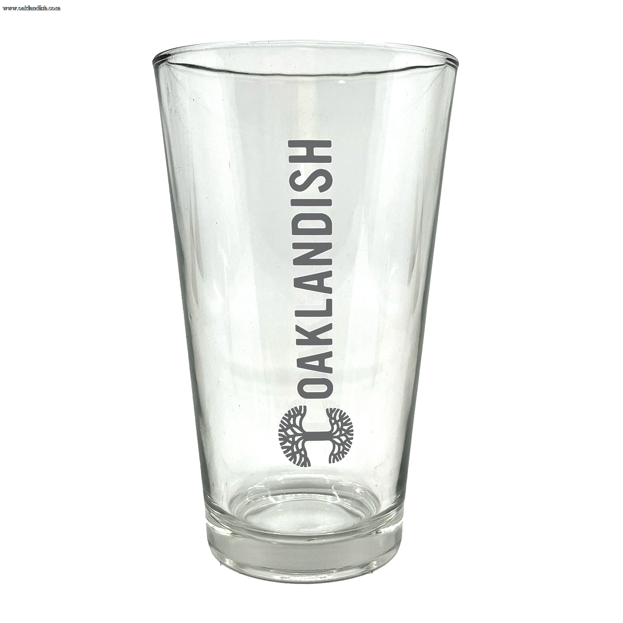 The Banner Pint Glass by Oaklandish is a clear, tall pint glass featuring a bold, dark grey Oaklandish logo in vertical text centered on one side. Below the text is a stylized black tree design within a circular outline. This piece of drinkware boasts a smooth finish and remains empty inside, awaiting your favorite beverage.