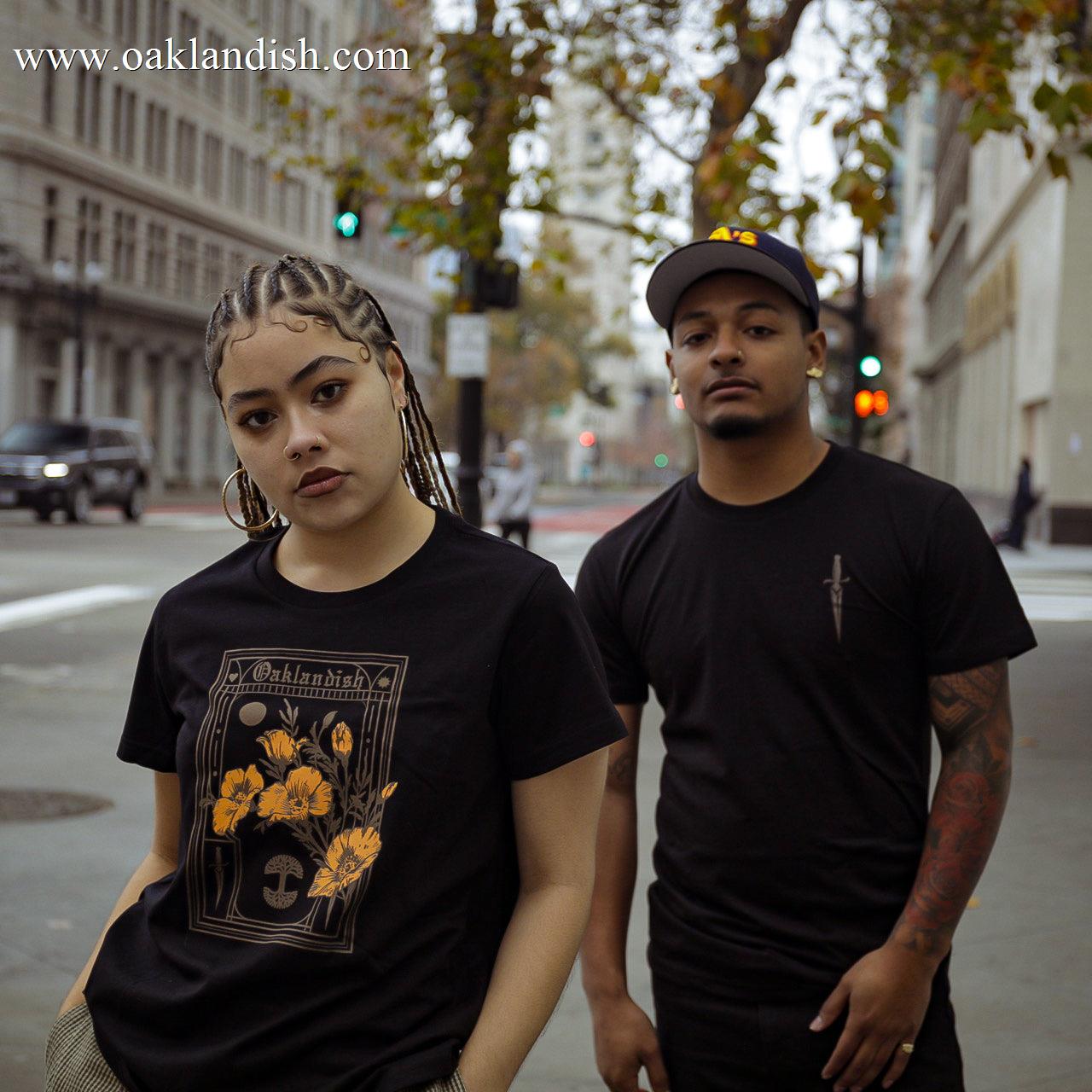 Two people stand in an urban street with buildings and trees behind them. The person on the left wears a black shirt with a floral and text design featuring poppies, has braided hair, and tilts their head slightly. The person on the right wears Oaklandish's Women's Blossom Tee, a cap, and has visible tattoos on their arms.
