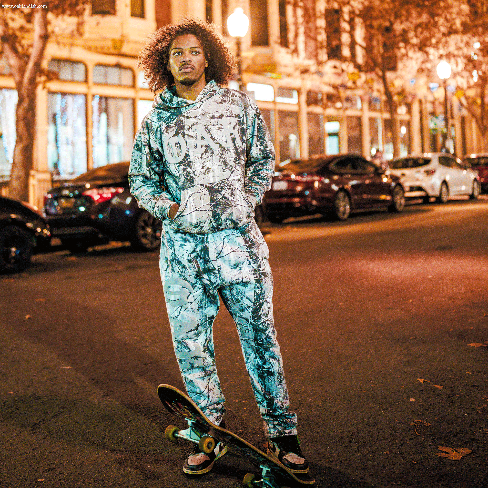 A person with curly hair stands on a street at night, wearing a matching Grey Wood Camo hoodie and Oaklandish Cork Pant. They hold a skateboard, hands in pockets, as cars line the brightly lit storefronts behind them.