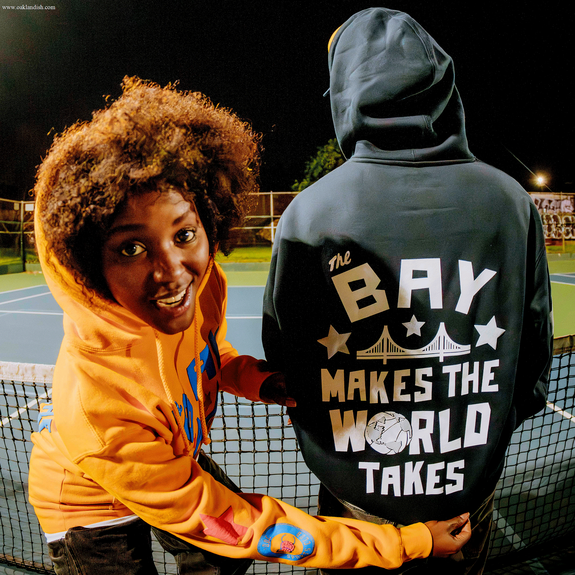 Two people are on a tennis court at night. One smiles at the camera in an orange hoodie. The other, showcasing local pride in Oaklandish’s Bay Bridge Hoodie by DOC, faces away, highlighting Bay Area-inspired style under the night sky.