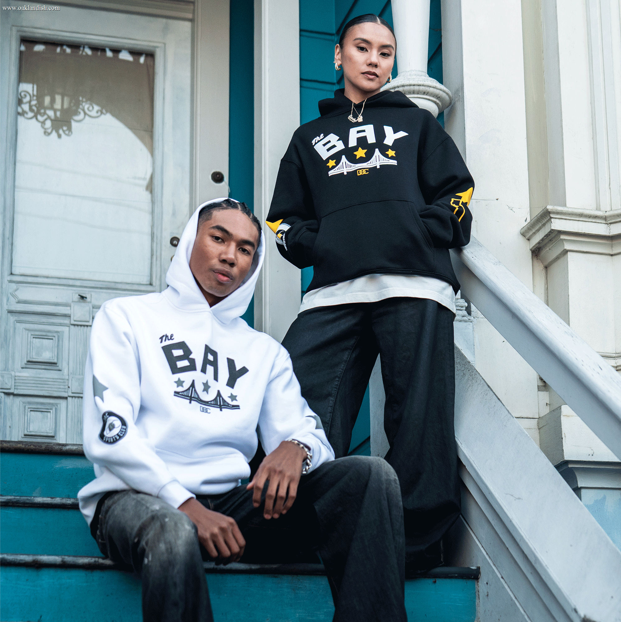 Two people pose on blue stairs, confidently wearing Oaklandish's Bay Bridge Hoodie By DOC V2—one in white, one in black—each with “THE BAY” and star graphics for a bold urban look.
