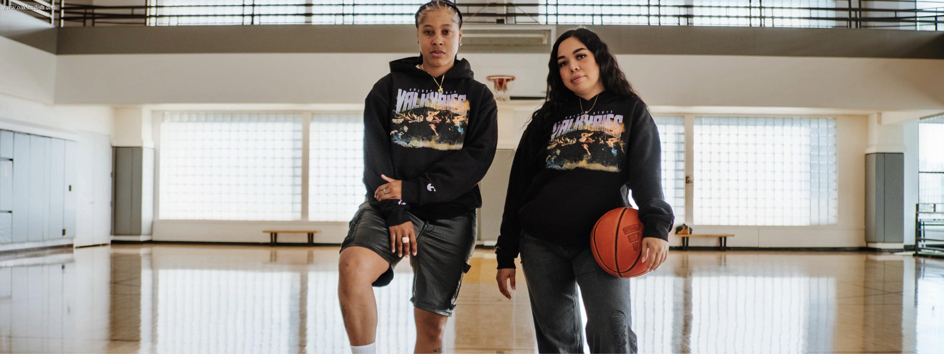 Two women in camo pants and “Valkyries” shirts stand confidently under a concrete overpass near a worn basketball hoop, smiling at the camera in natural daylight.