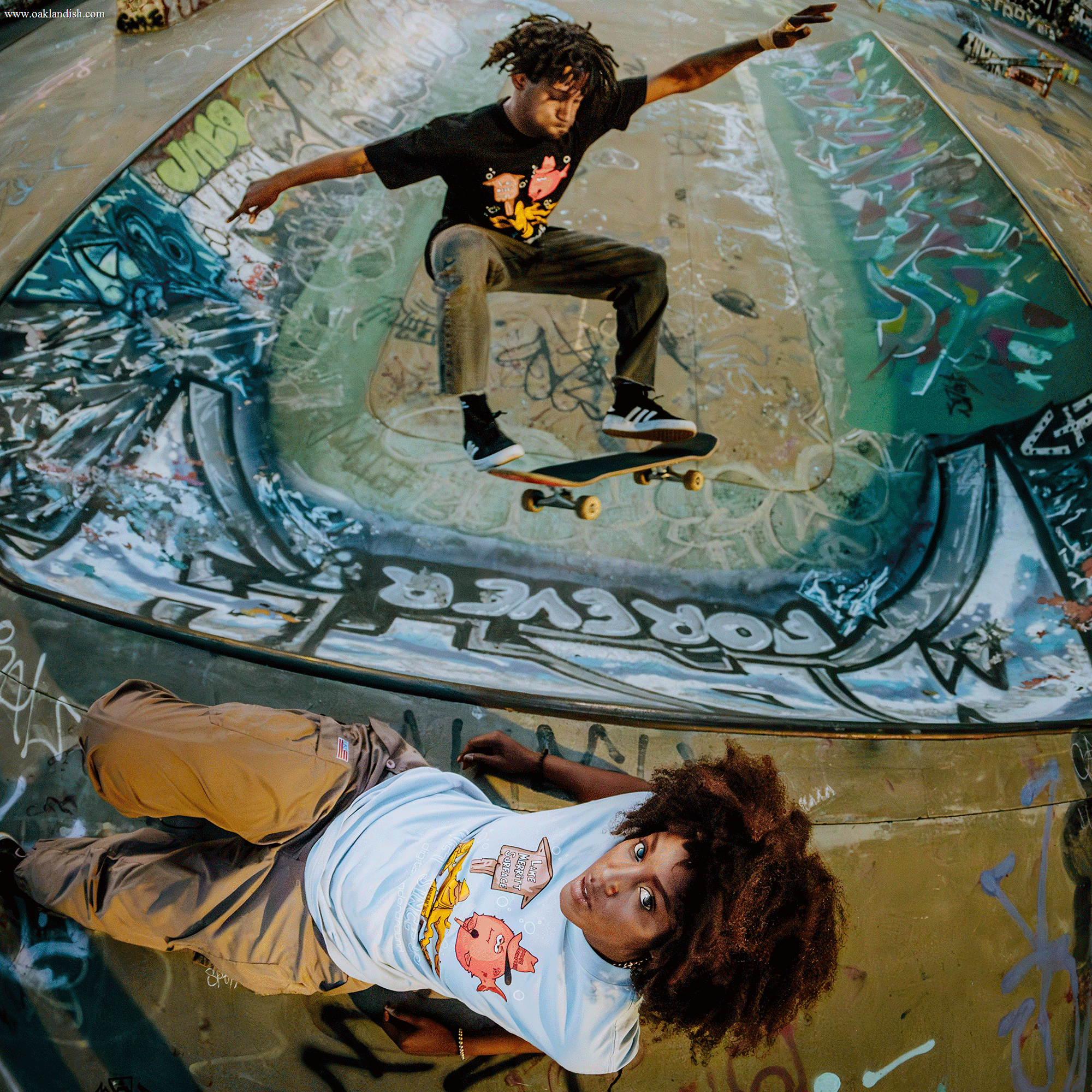 A skateboarder performs a trick above an Oakland roots-themed graffiti-covered skate bowl. Nearby, a curly-haired person lies on the ground, gazing up at the camera. Both wear Oaklandish's Wet Since '88 Tees with cartoon-like graphics, creating a vibrant and dynamic scene full of street art.
