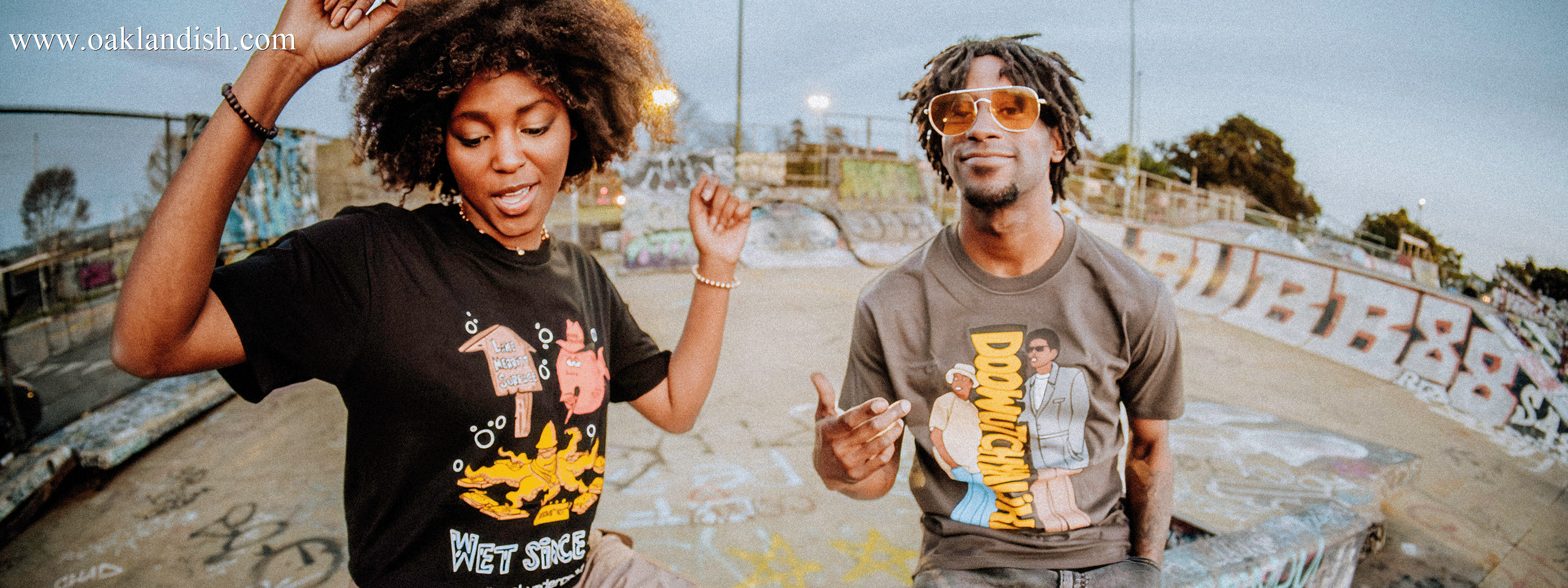 Two people dancing at a graffiti-covered skate park. They wear graphic t-shirts and have joyful expressions. The woman has curly hair and the man wears sunglasses. Skaters are visible in the background.