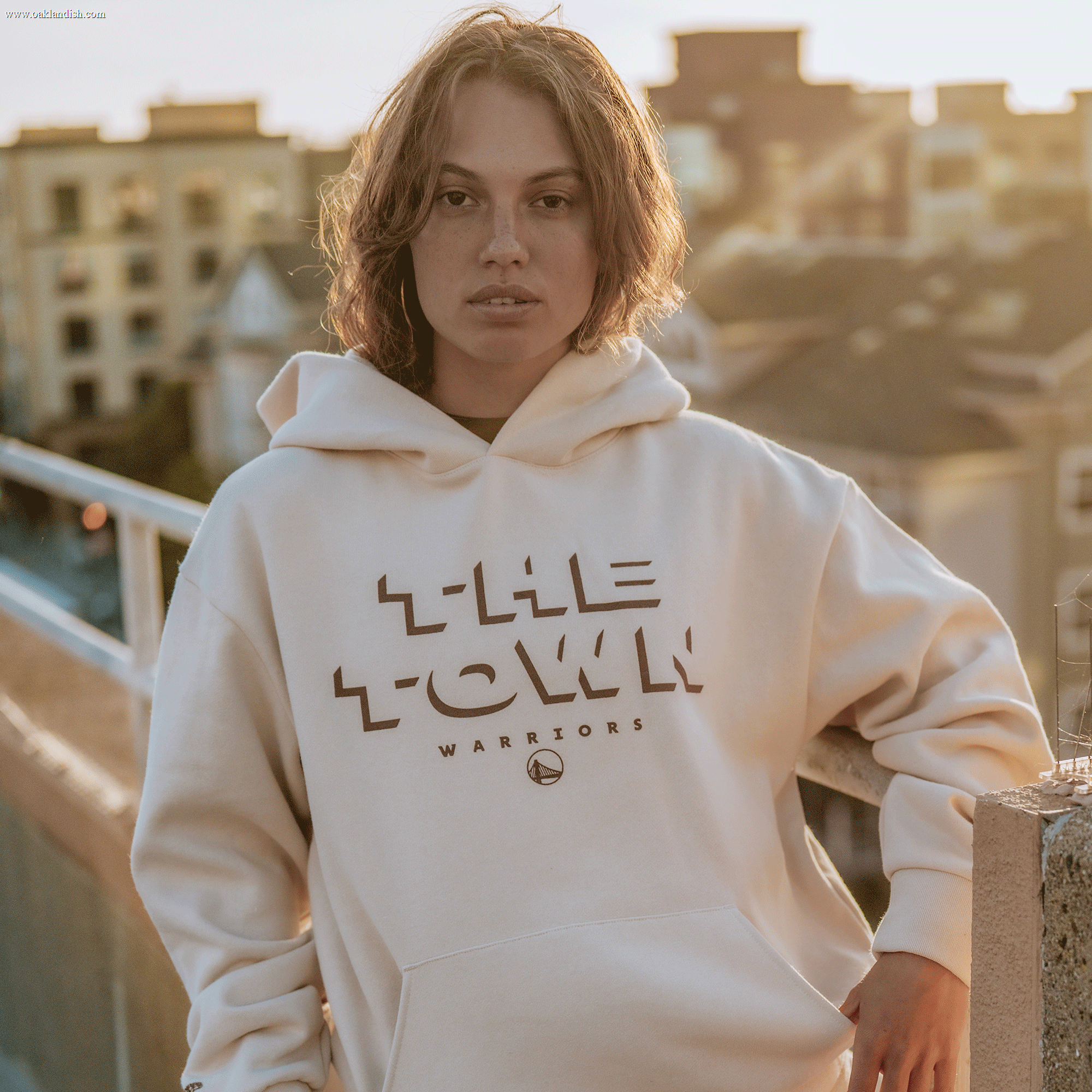 A person with short brown hair, wearing a white Ghost Block Letters Hoodie by Other, stands on a rooftop at sunset. Apartment buildings and a blurred cityscape are in the background, illuminated by warm, soft light.