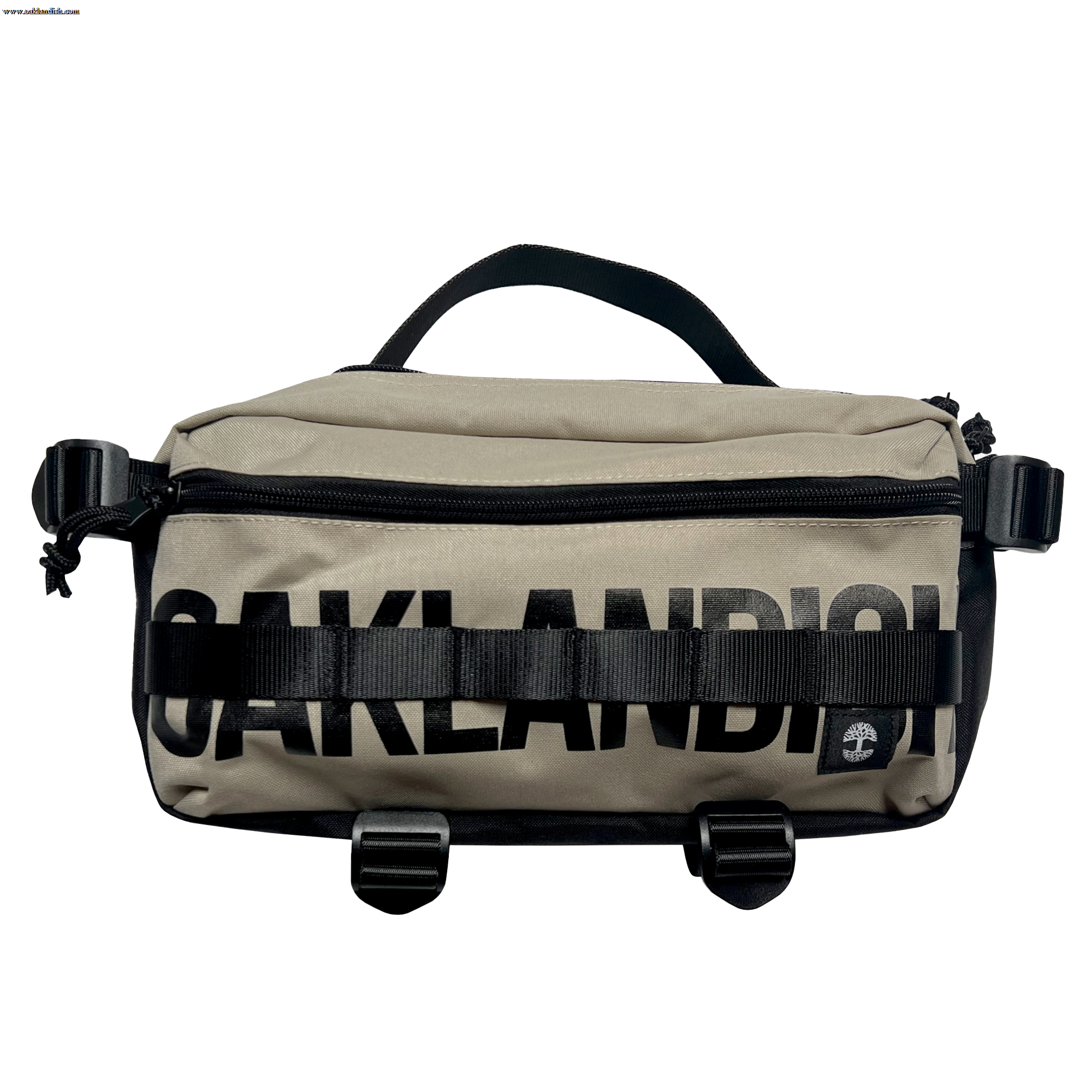 The Oaklandish Hip Bag is a beige and black unisex sling pack with "OAKLANDISH" boldly printed across the front. It features a black handle, buckles, and straps, along with a small circular logo patch on the lower right corner.