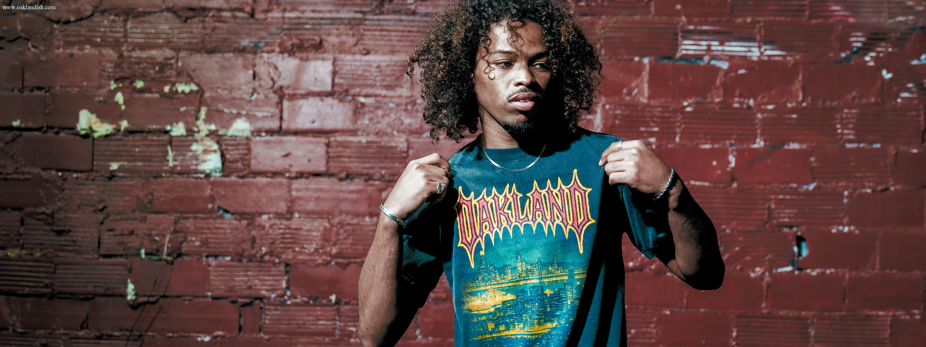 A person with curly hair stands outdoors wearing a white T-shirt with a nature-themed design. The background features trees and a sunset, creating a warm, serene atmosphere.