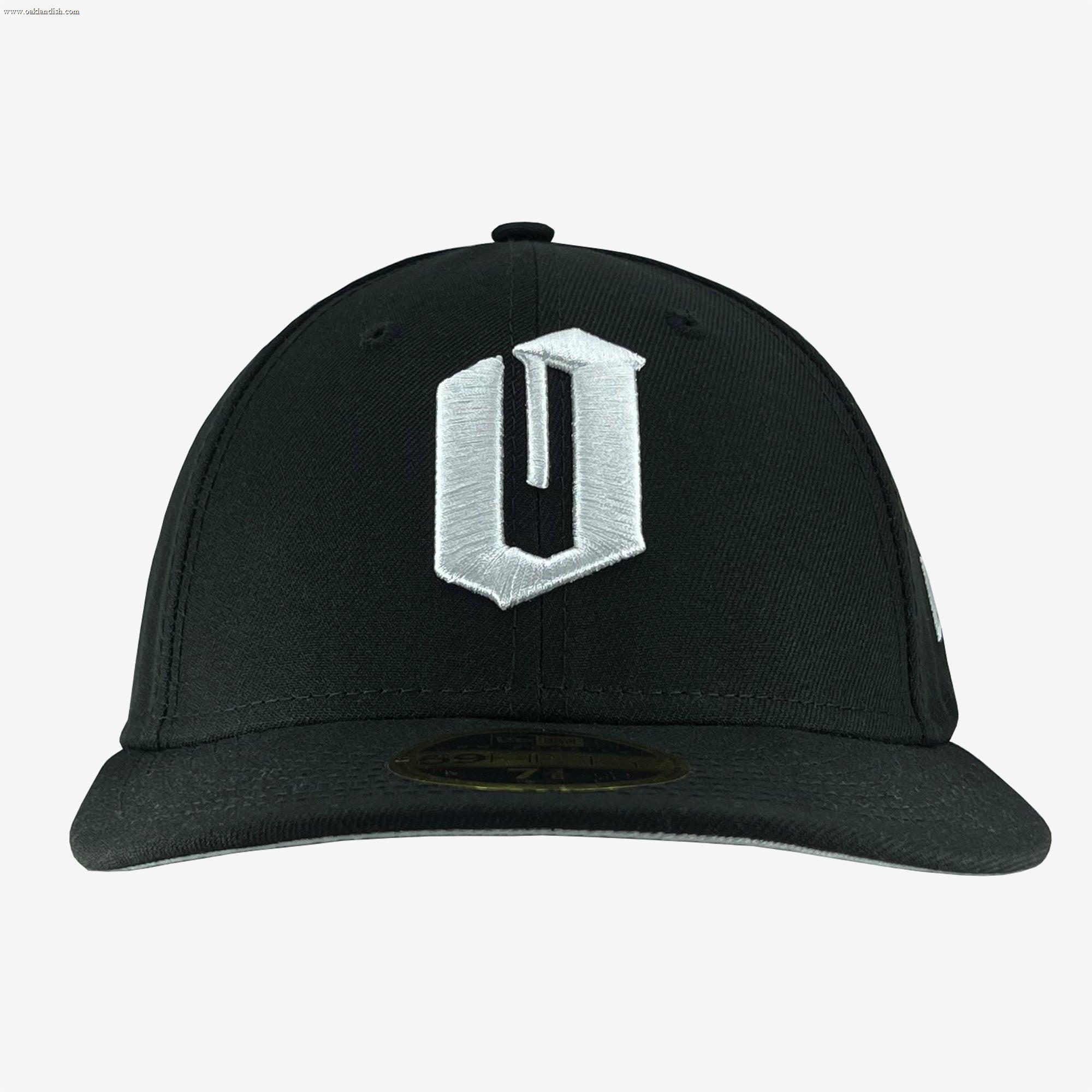 A black New Era Official Low Profile 59FIFTY Fitted Cap by Oaklandish, with a prominent white letter "U" embroidered on the front panel. This cap features a flat brim and includes a sticker visible on the underside of the brim. The background is plain white.
