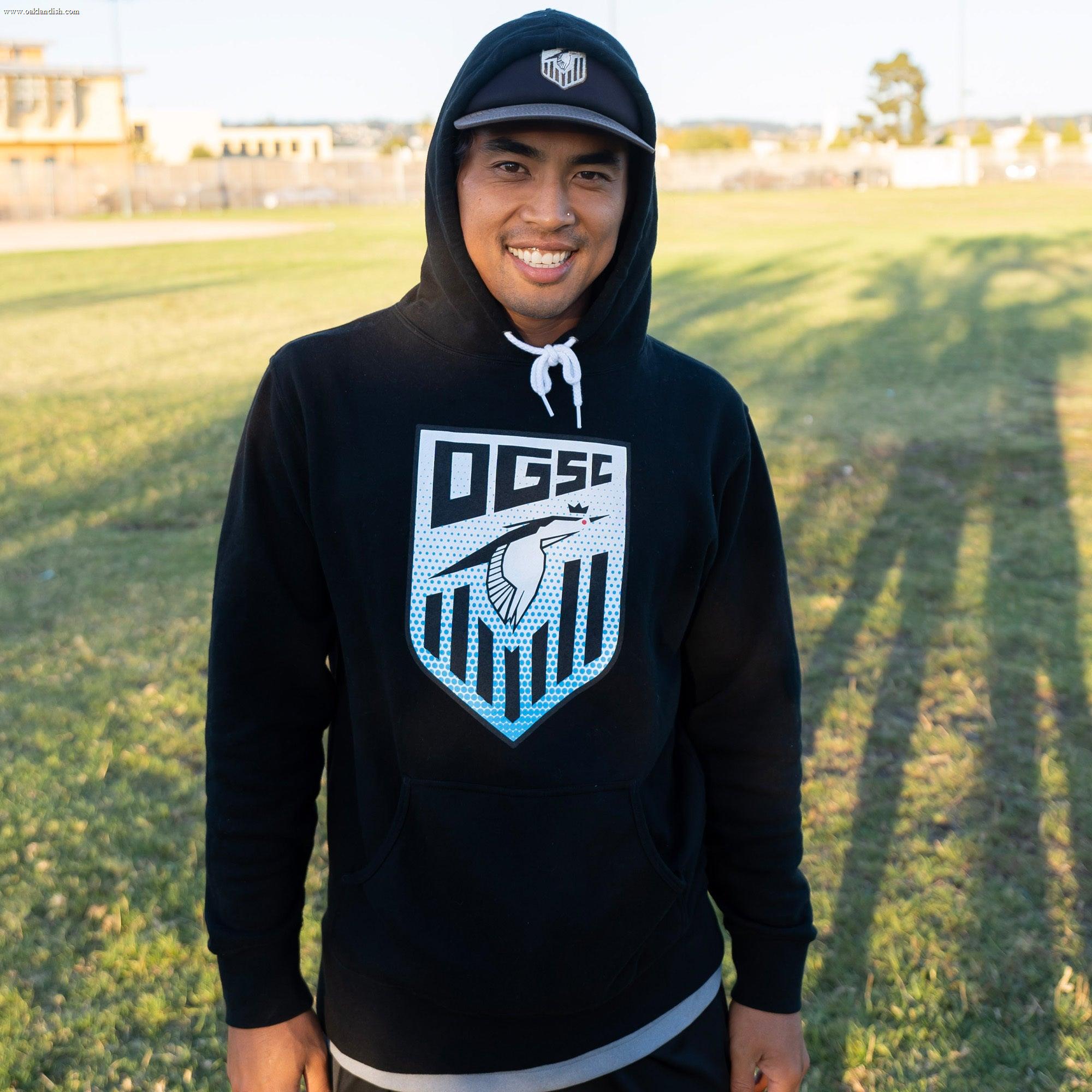 A person wearing a black Partner Oakland Genesis Heron Hoodie stands on a grassy field, smiling and donning a matching cap. In the background, buildings and trees cast shadows on the sunny setting. This nonprofit organization supports youth soccer in the community.