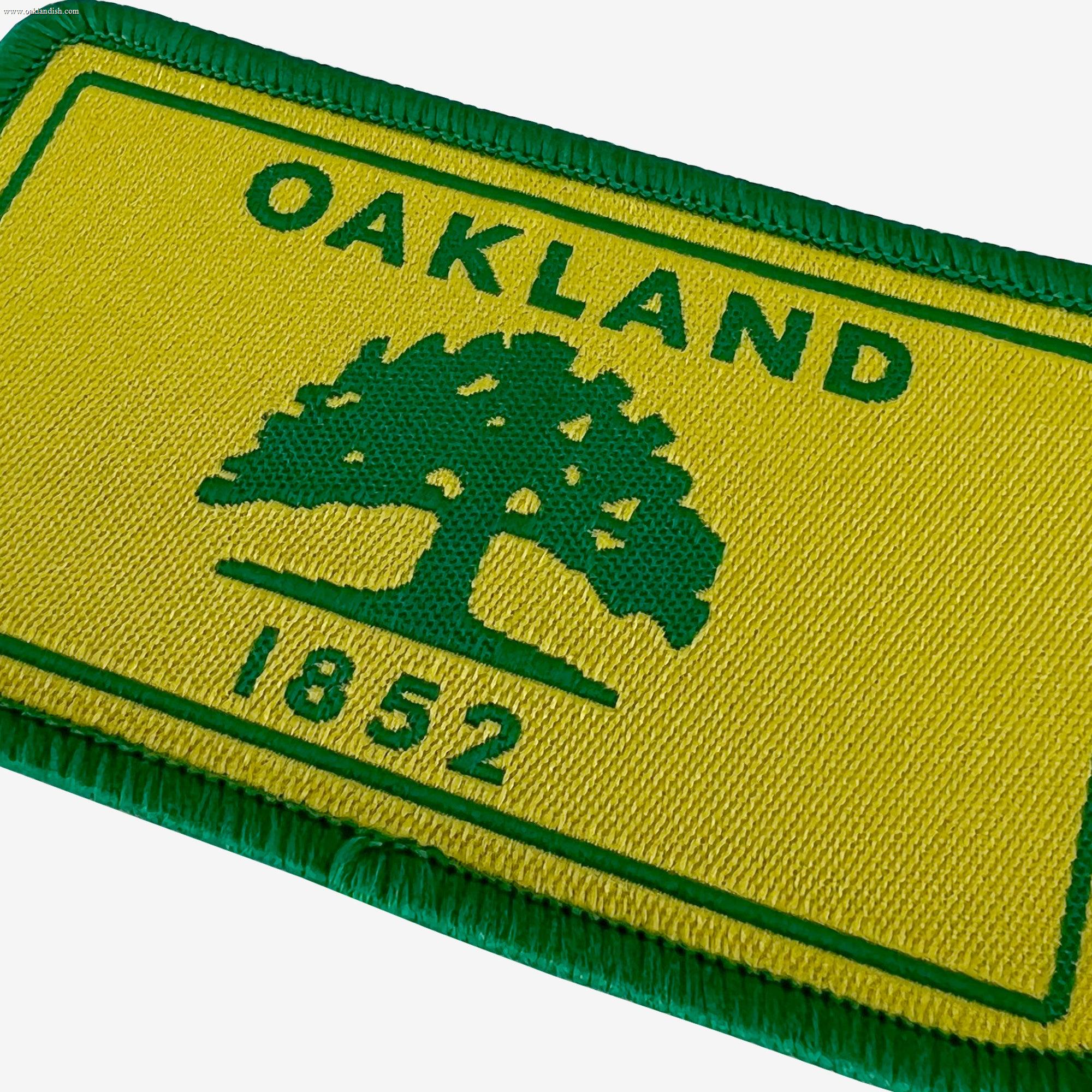 The Oaklandish Oakland Flag Patch is a rectangular, embroidered iron-on decoration with a green border and yellow background. It features the word "OAKLAND" in green at the top, a green silhouette of a tree below that, and the number "1852" in green at the bottom. It's perfect for adding flair to your pillowcase using a hot iron.