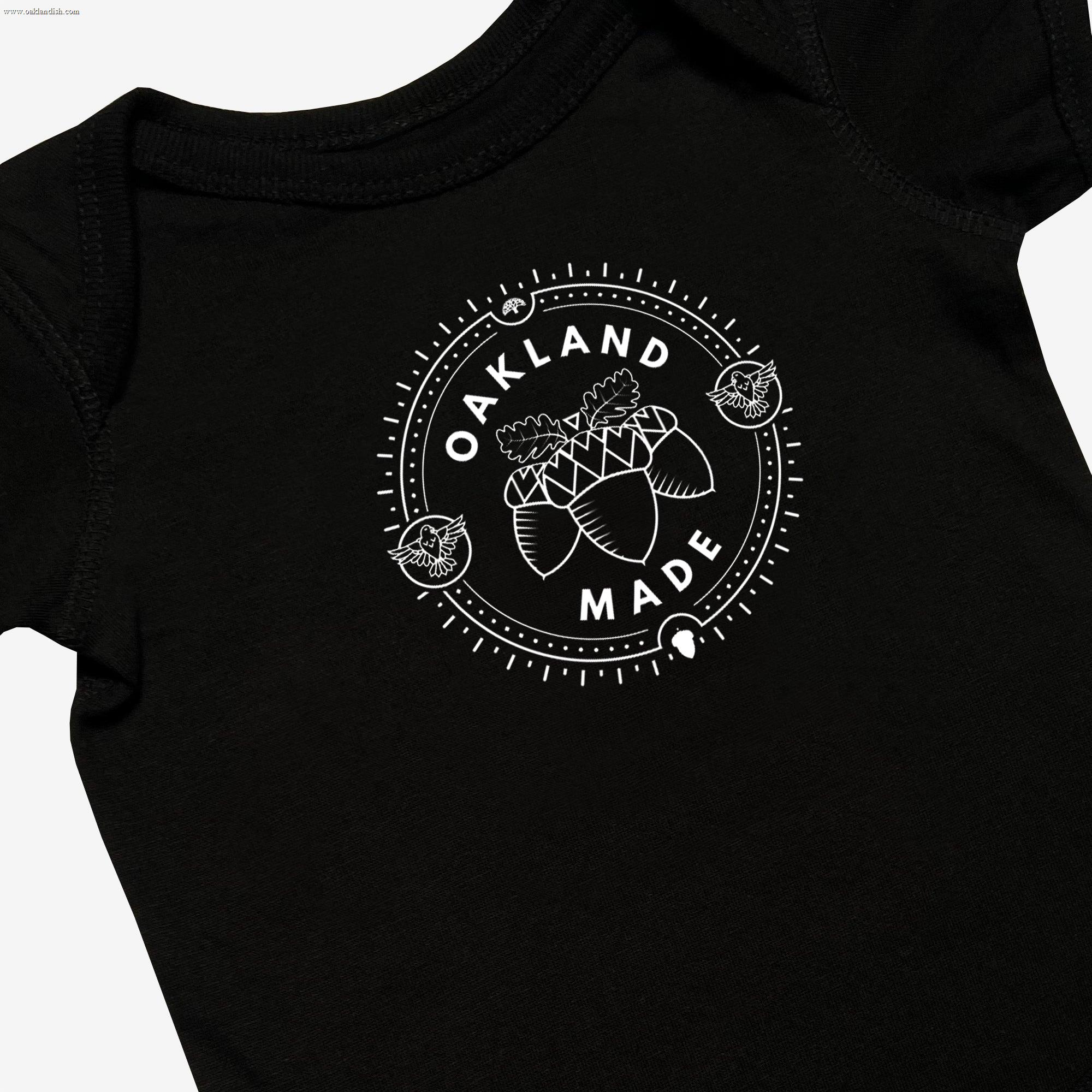 Close-up of black infant one-piece with a white circle graphic with the words “Oakland Made” and hand-drawn chestnuts.