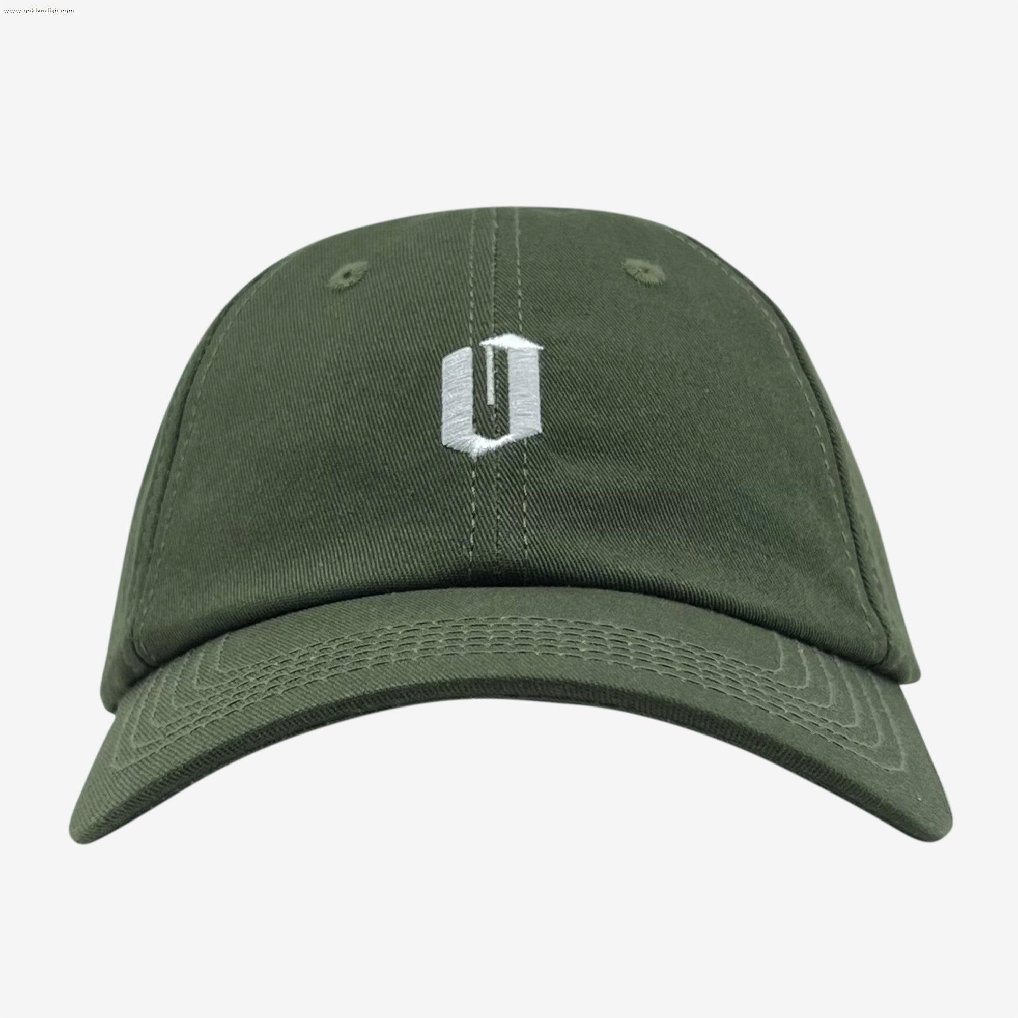 The Oaklandish Official Micro Logo Dad Hat in green is displayed against a plain white background. The cap prominently features a small white embroidered "U" logo on the front, just above the bill. Its design is minimalist, showcasing a curved brim, visible stitching along the panels and edges, and an adjustable strap for an ideal fit.