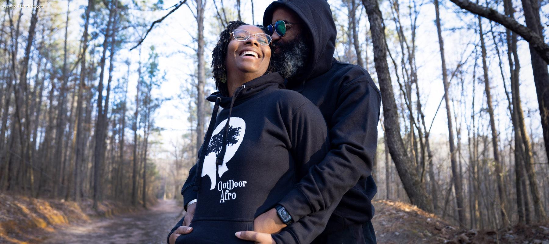 A smiling couple in black hoodies stands embraced on a forest path, surrounded by tall trees. The front person’s hoodie reads "Outdoor Afro" with an afro silhouette and tree graphic.