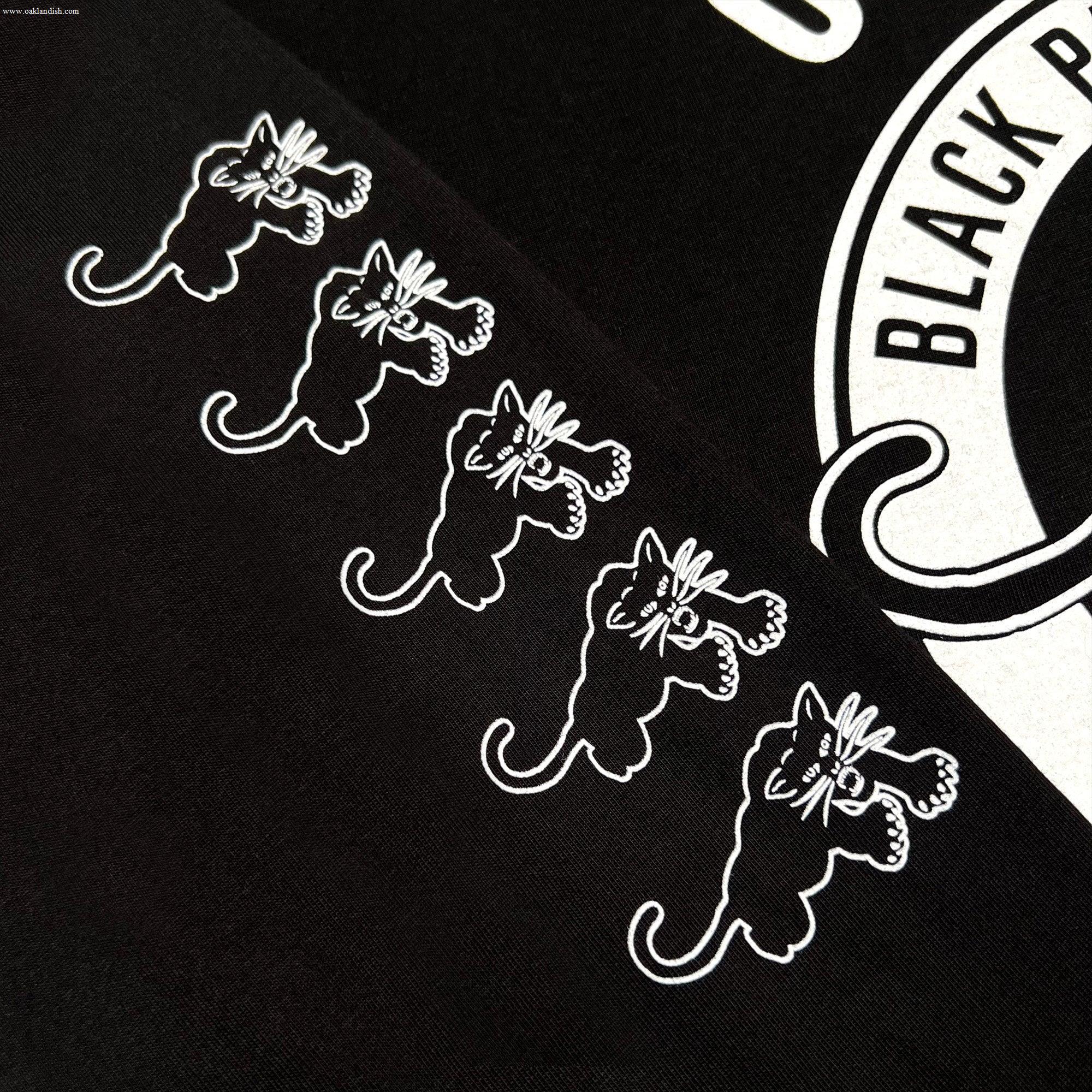The image features the Black Panther Power LS Tee by Oaklandish, showcasing a black fabric adorned with five identical white-printed cat illustrations aligned horizontally. The cats exhibit playful, wide-eyed expressions and raised paws. To the right, there's a partial view of an ornate white circular logo, reflecting the bold legacy of Black Panther Power.