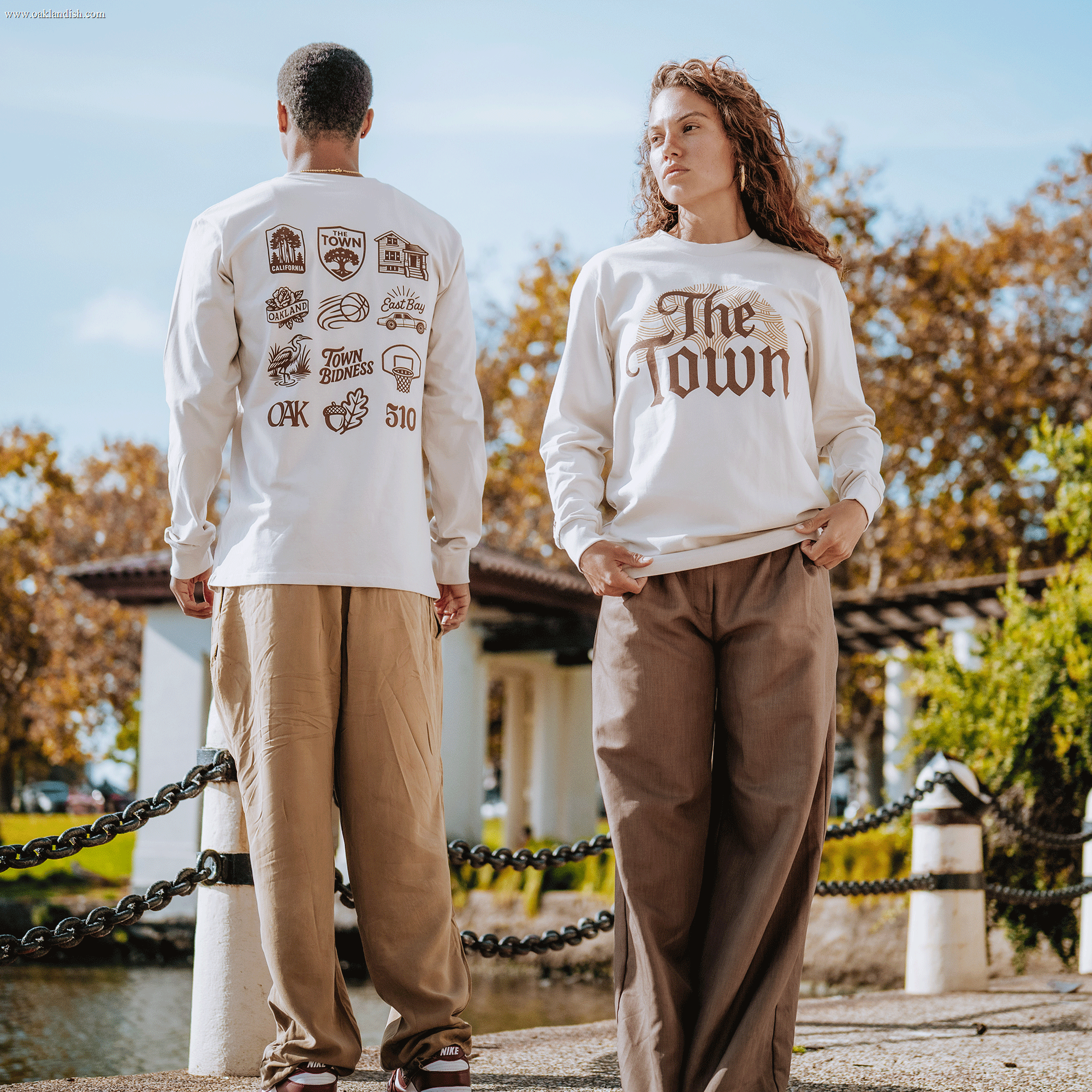 Two models stand outdoors near the water. The man, facing away, wears Other’s Pirate Black-Letter LS Tee in cream from the Warriors x Oaklandish Town Collection, featuring Oakland-themed graphics on the back. The woman faces forward in a matching cream Pirate Black-Letter LS Tee that reads "The Town" in brown script overlaid on topographical lines. Both pair their tops with loose-fitting, earth-tone pants.