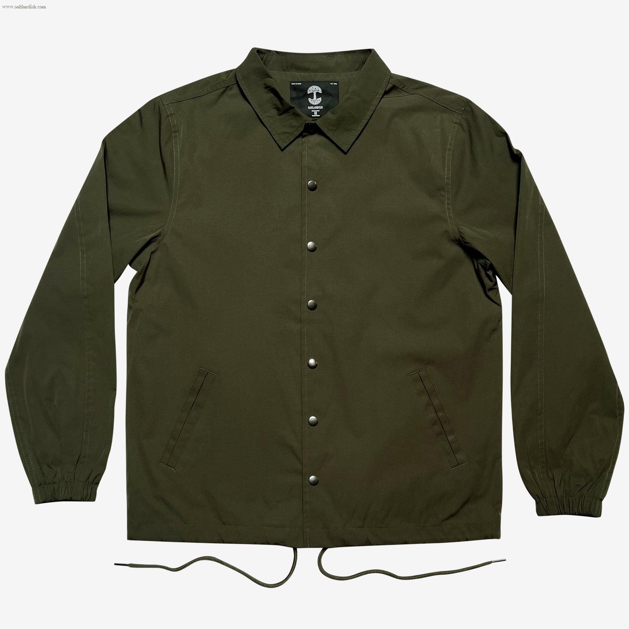 This image showcases the Premium Coaches Jacket by Oaklandish in olive green, featuring long sleeves and a pointed collar. The front displays a row of metallic snap buttons and two diagonal pockets. The elastic cuffs and dual drawstrings at the bottom hem add functionality to this water-resistant outerwear, which exudes a sporty and casual style.