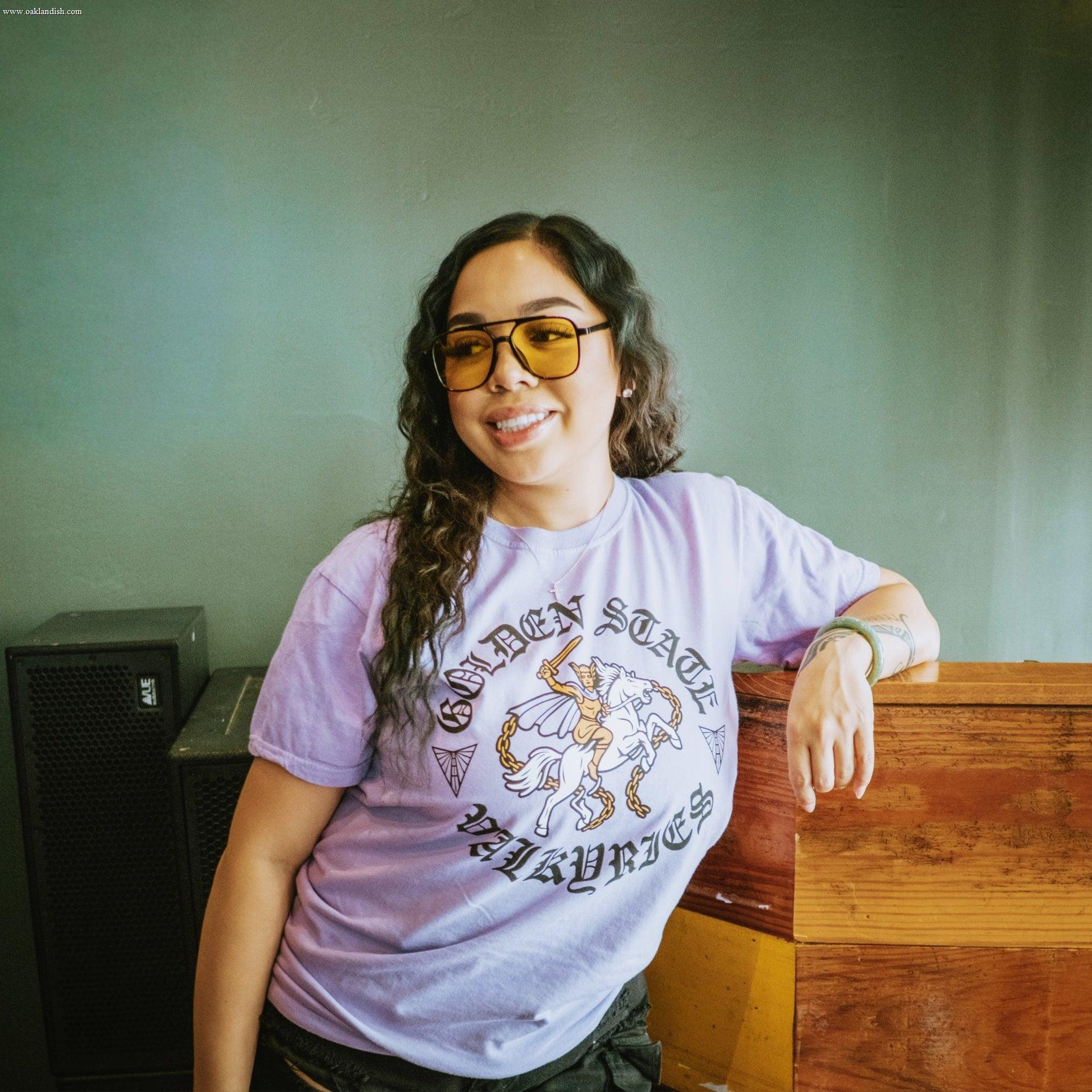 A woman with long wavy hair, yellow-tinted sunglasses, and a lavender GSV ROTV Tee smiles as she leans on a wooden surface. A gray wall and black speaker appear in the background.