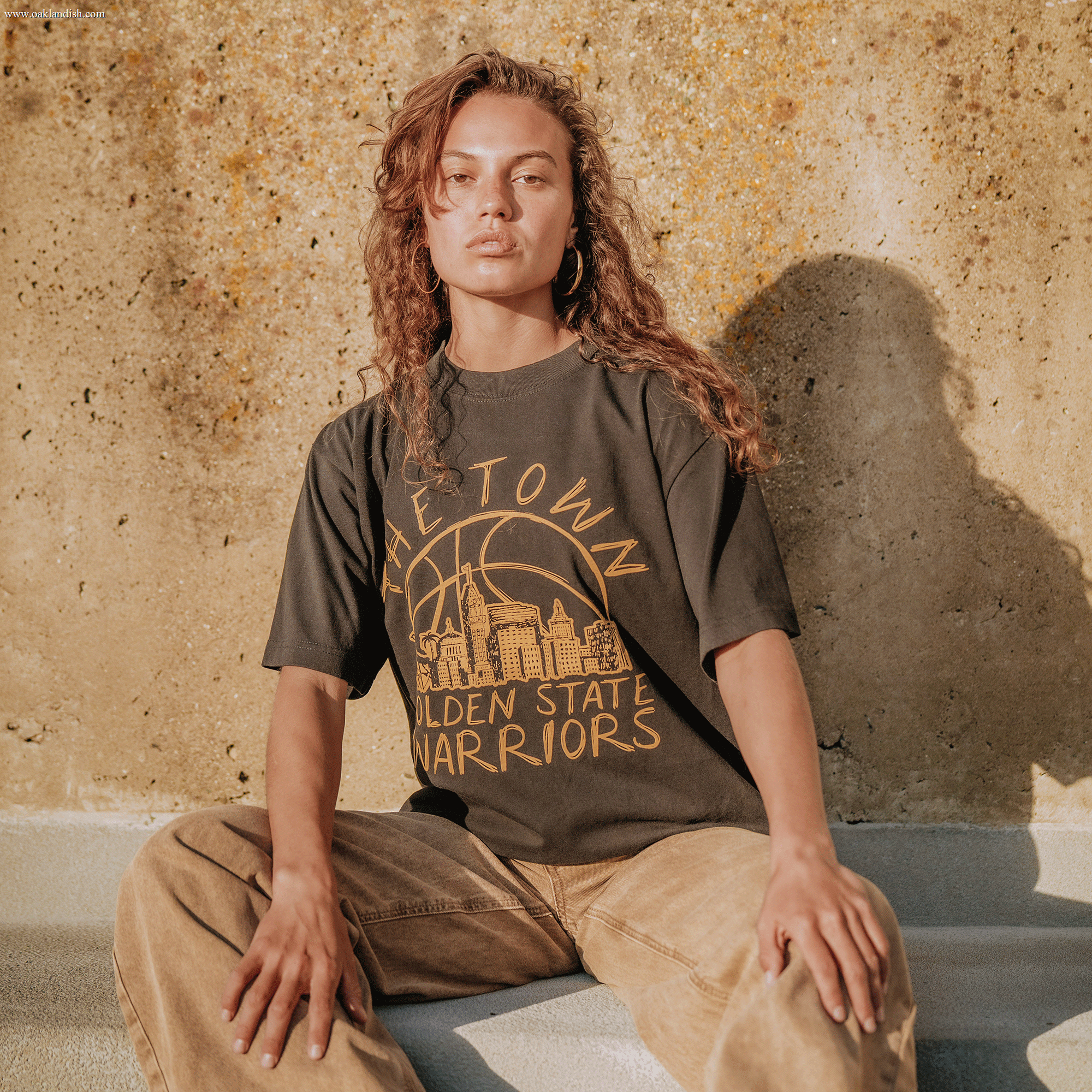 A woman with curly hair sits on concrete steps in the sunlight, leaning against a textured wall. She wears the Other brand's Town Doodle Tee, a black t-shirt from the Town Collection featuring a yellow city graphic, paired with light brown pants, and looks confidently at the camera.
