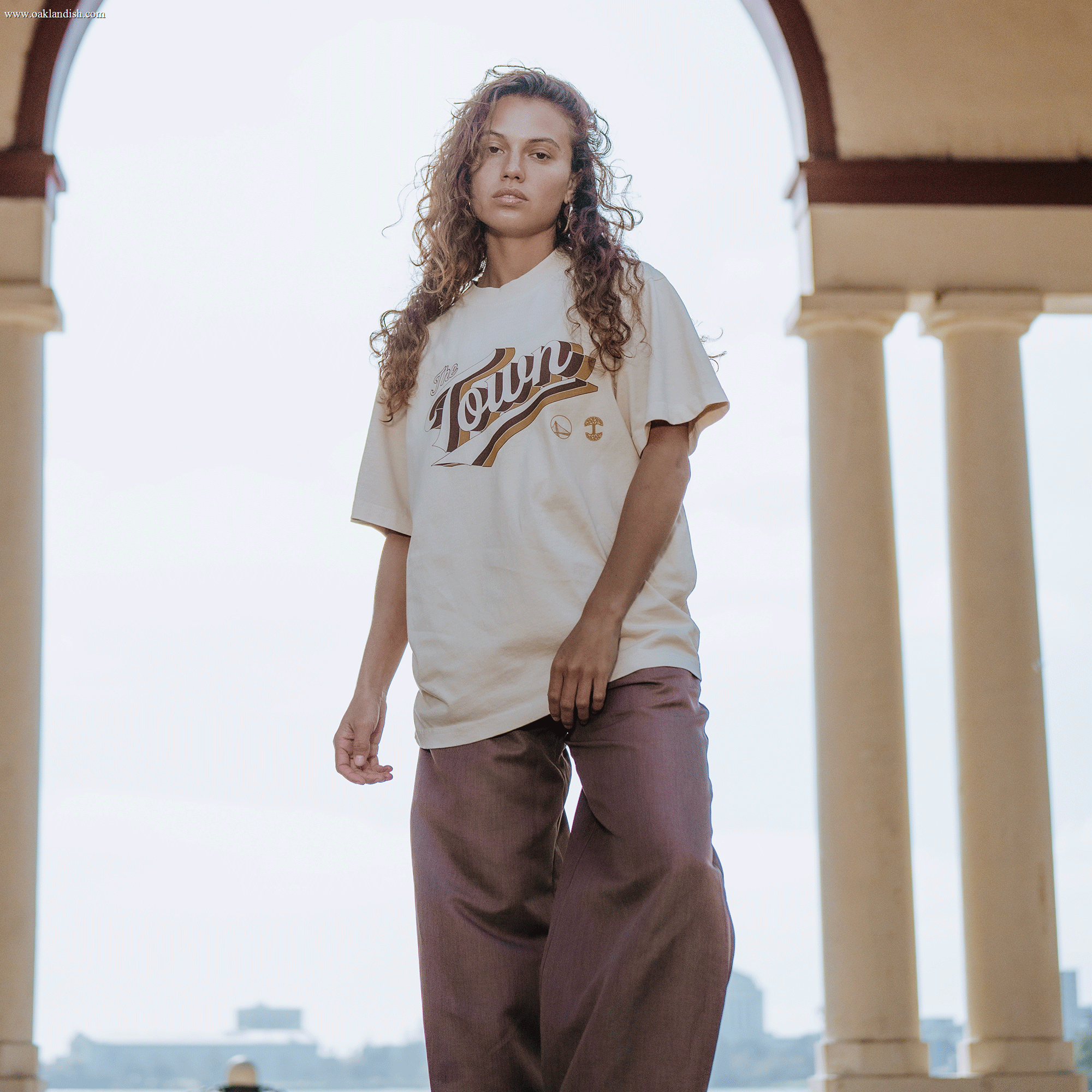 A person with curly hair stands beneath an arched structure, confidently gazing at the camera. They wear the cream Vintage Swash Tee from Other and loose brown pants. The backdrop features bright, cloudy skies and blurred buildings.
