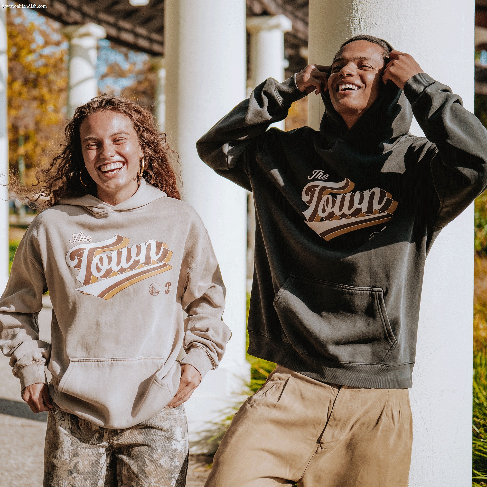 Two people stand outside under a pergola, smiling and laughing. Both wear the Vintage Swash Hoodie by Other—one in tan and the other in dark gray—each featuring “The Town” in stylized script from the Warriors x Oaklandish collaboration. Sunlight creates a warm, cheerful atmosphere with greenery and white columns in the background.