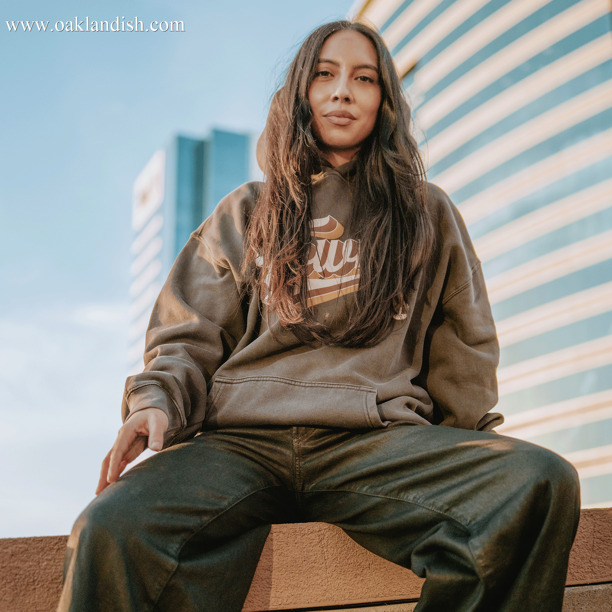 A woman with long, dark hair sits on a ledge, wearing the Other Vintage Swash Hoodie in brown and dark pants. Photographed from a low angle, she gazes toward the camera with a neutral expression. In the background, a modern glass-clad skyscraper reflects the bright, golden late-afternoon sky.