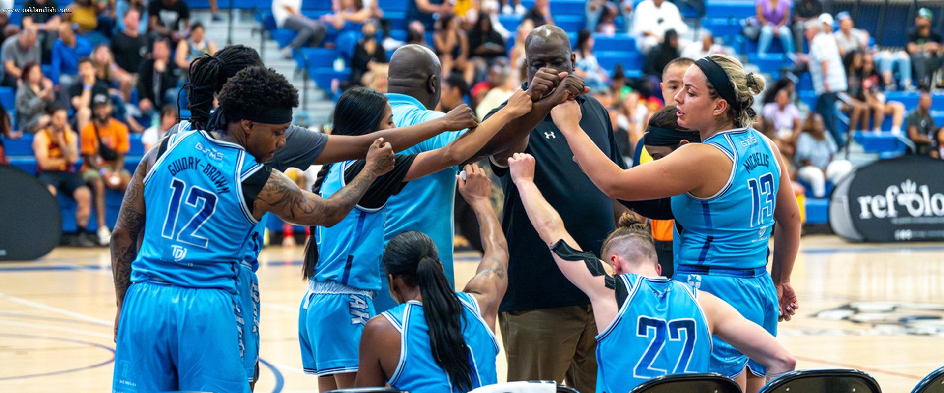 A basketball team in blue jerseys huddles with their coach, arms raised in unity, on a gym court with a crowd of spectators in the bleachers behind them.