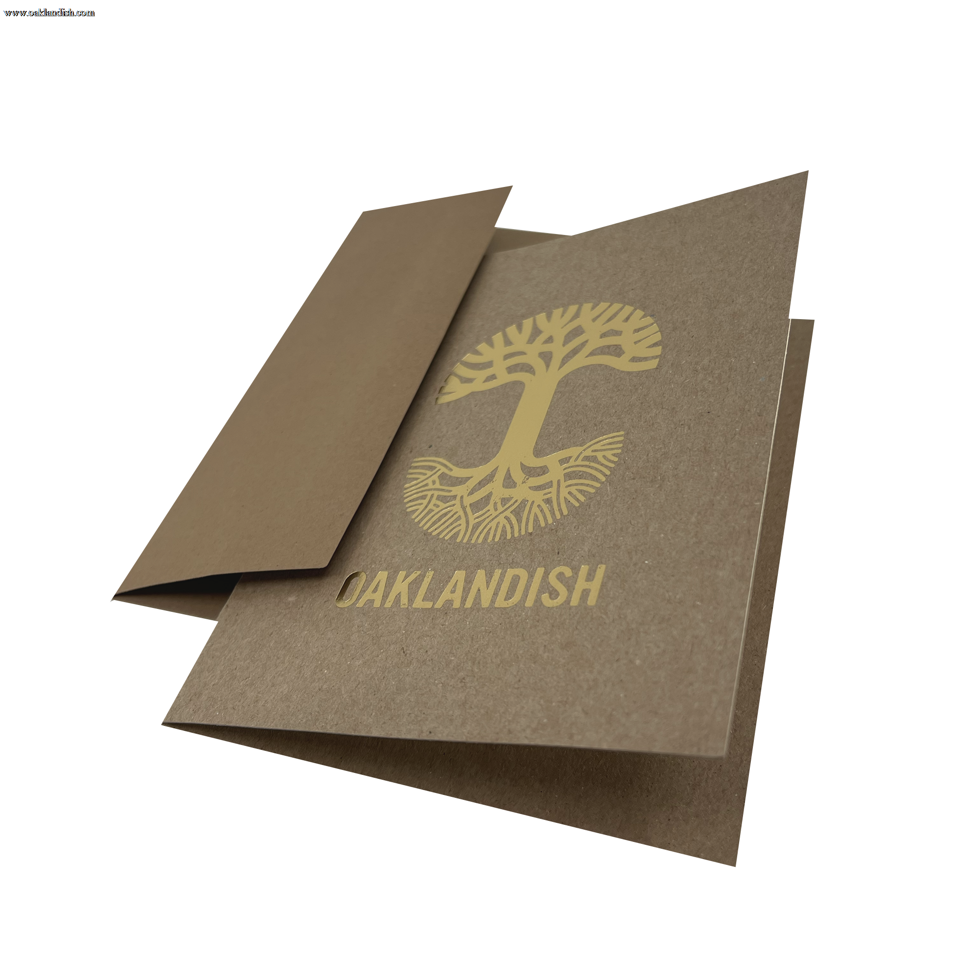 An "Oaklandish New Logo Card" by Never Elsewhere, crafted from recycled brown kraft paper, lies partially open on a white background. The card displays a stylized gold tree with exposed roots and the word "OAKLANDISH" printed beneath it using letterpress print. One flap is partially folded inward, revealing the inside of the card.