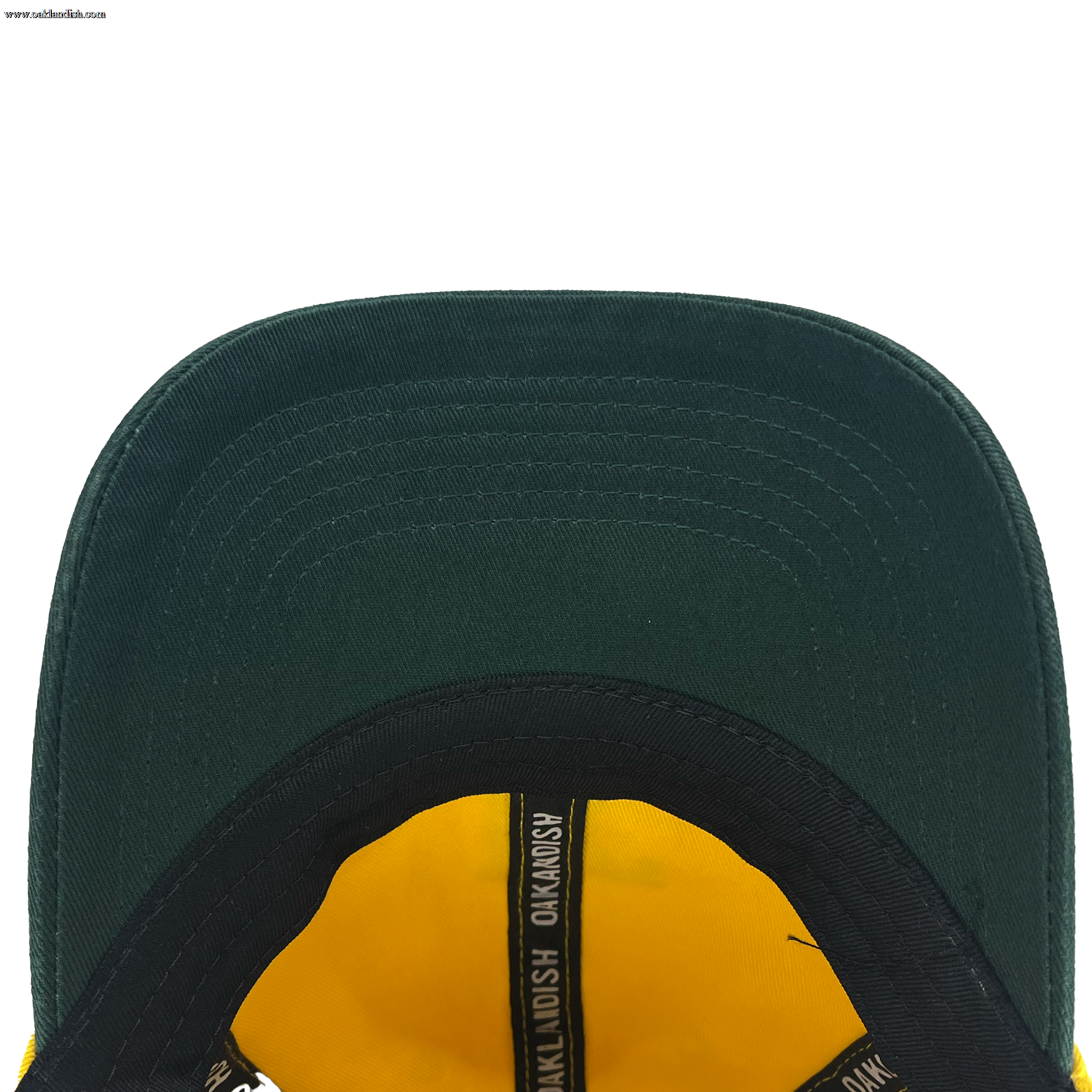 An overhead view of the underside of the Oakland Ballers Dad Hat reveals a dark green baseball cap. The brim stands out, adorned with multiple stitched lines. Inside, there is yellow fabric and a black band featuring the repeated text "OAKMOSH," representing Oakland Ballers from the Pioneer League.