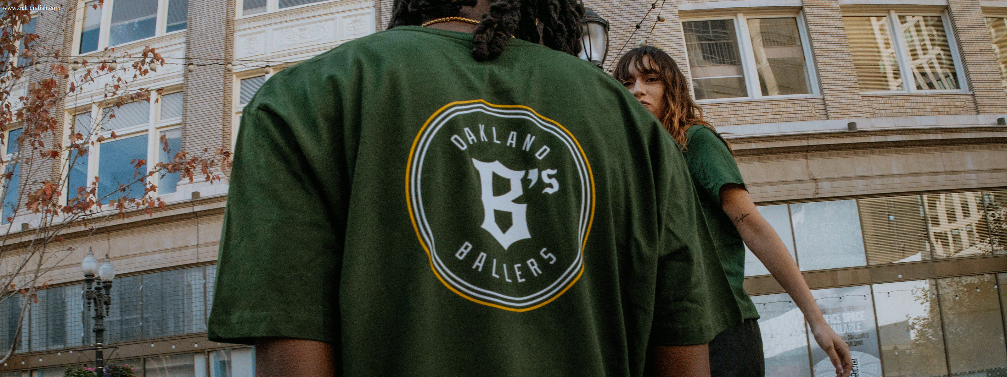 Two people stand outside in front of a building; the person in the foreground wears a green shirt with a circular logo reading "Oakland Ballers B’s," while the other looks toward the camera.