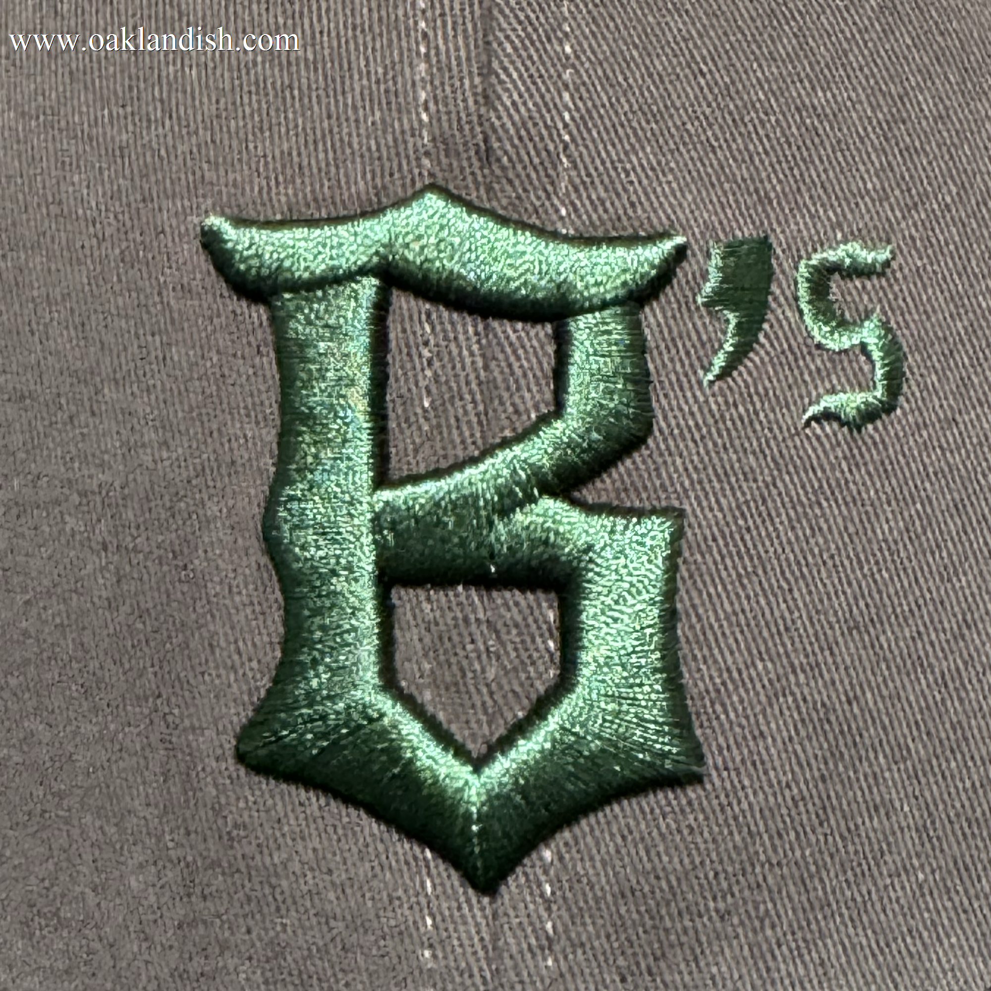A close-up of the Oakland Ballers Logo Hat by Oakland Ballers shows gray fabric with a large dark green embroidered gothic "B," plus an apostrophe and "S," representing the new Oakland Pioneer League baseball team’s logo.