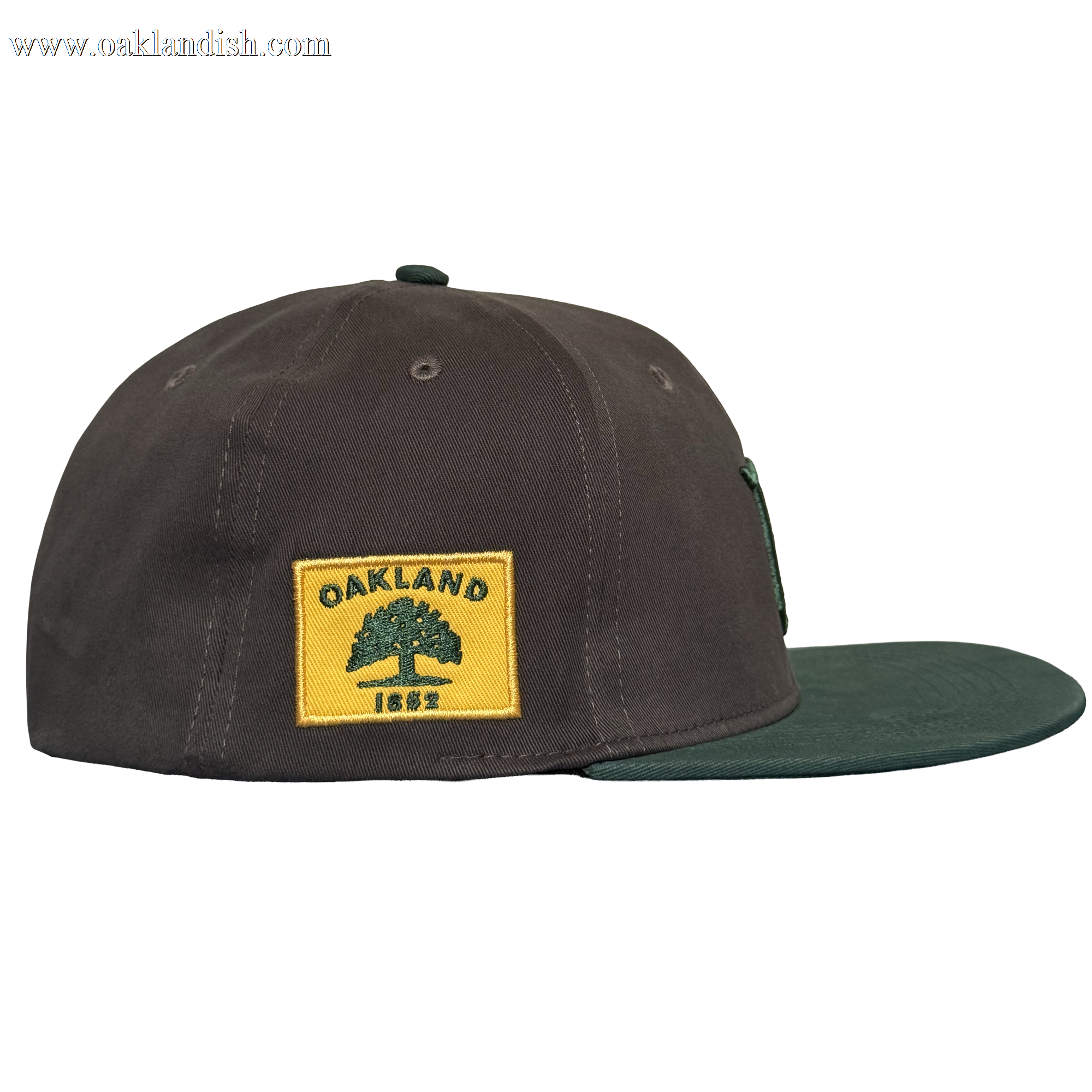 The Oakland Ballers Logo Hat, by Oakland Ballers, is a dark brown cap with a green bill, shown in side view. A yellow patch on the side displays a green tree, “OAKLAND,” and “1852.” The background is plain white.