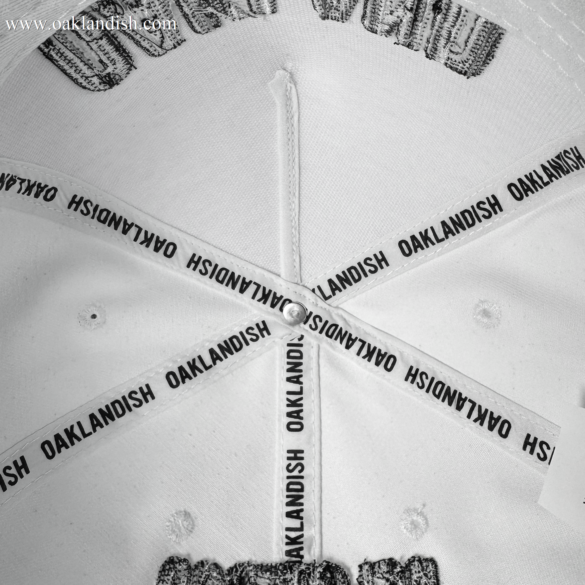 Close-up of the inside top of the white Oakland Beisbol 5 Panel Cap by Oakland Ballers. Four intersecting fabric strips, each printed with "OAKLANDISH" in black, plus white stitching and grommets evoke classic baseball team style.