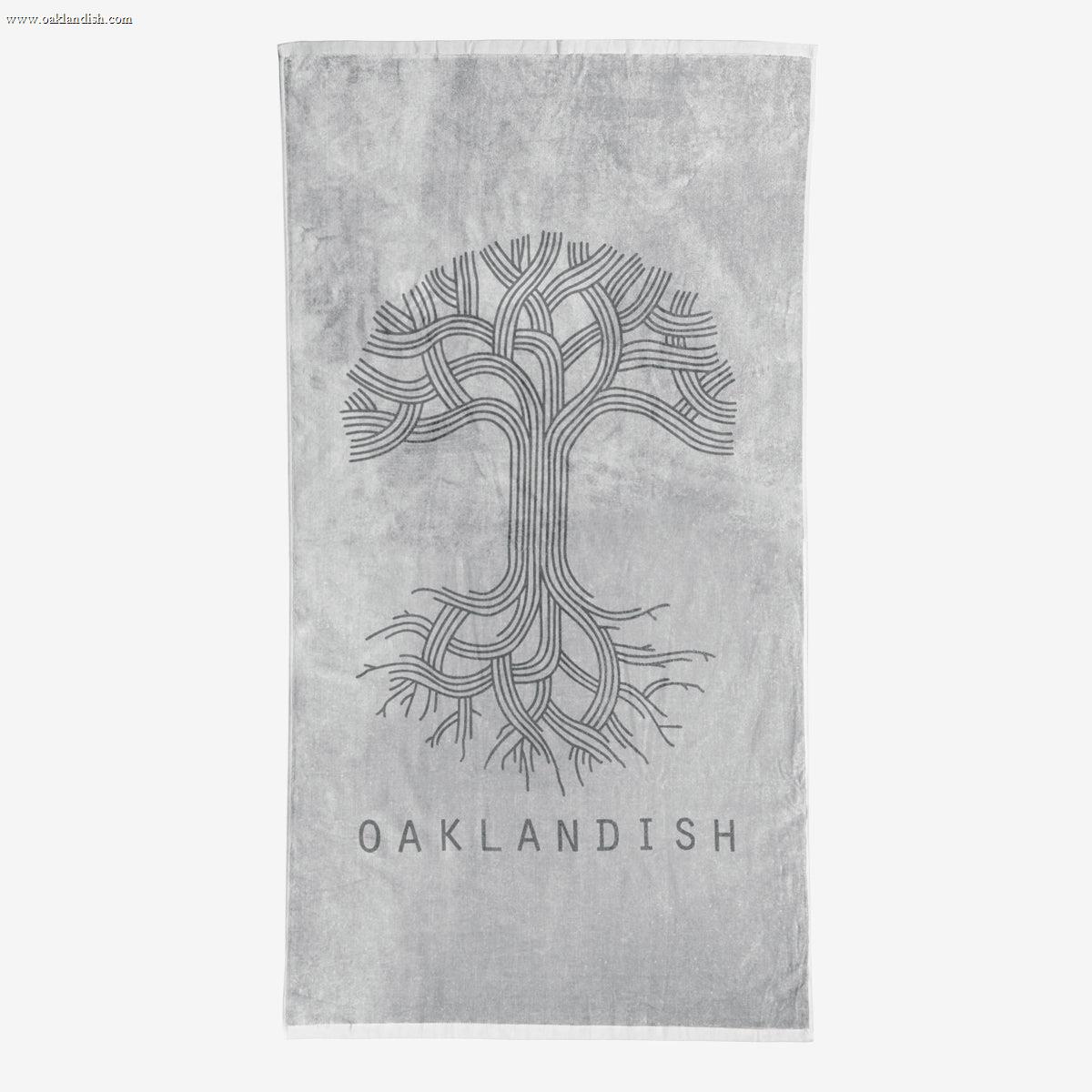 The Classic Logo Beach Towel by Oaklandish is a grey, oversized towel featuring an intricate, stylized tree design with numerous curved branches and roots in the center. Below the tree, "OAKLANDISH" is prominently displayed in capital letters. Made from 100% ringspun cotton, this super soft towel features a lighter grey background.