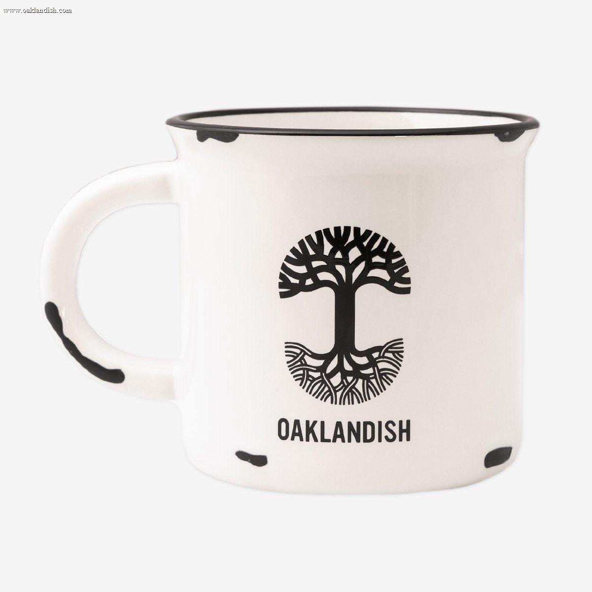 The 15 oz Ventura Mug by Oaklandish features a white ceramic body accented with a black rim and black speckles. The standout design includes a black tree logo with roots extending below it, accompanied by the "OAKLANDISH" text beneath, celebrating Oakland city pride. The handle is thoughtfully designed with matching speckles to complete the cohesive look.