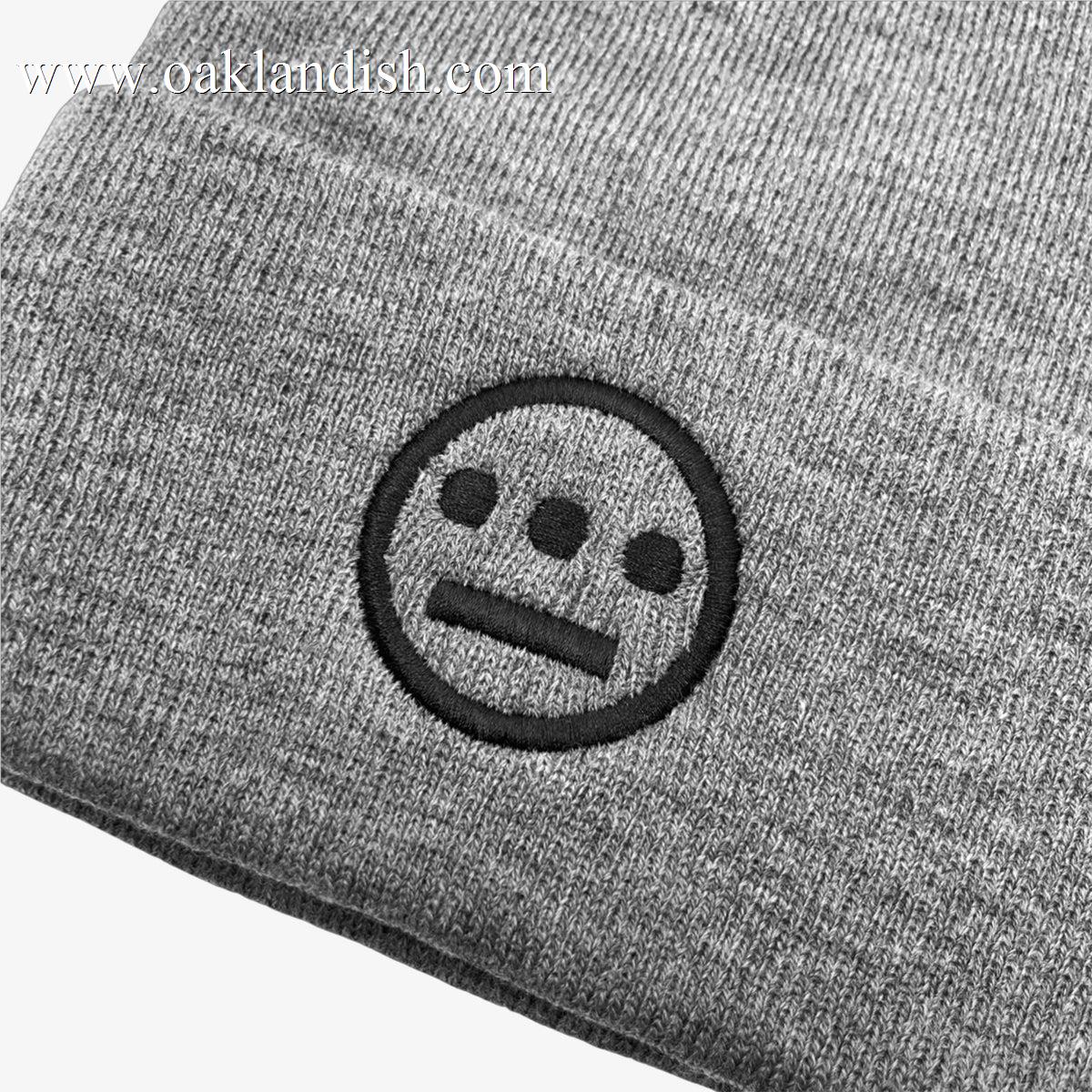Close-up of the Hiero Classic Cuff Beanie by Hieroglyphics featuring an embroidered logo. The logo showcases a simple, geometric face design reminiscent of hieroglyphics, with three horizontal dots forming the eyes and nose, and a horizontal line for the mouth, all stitched in black thread on a gray knit fabric.