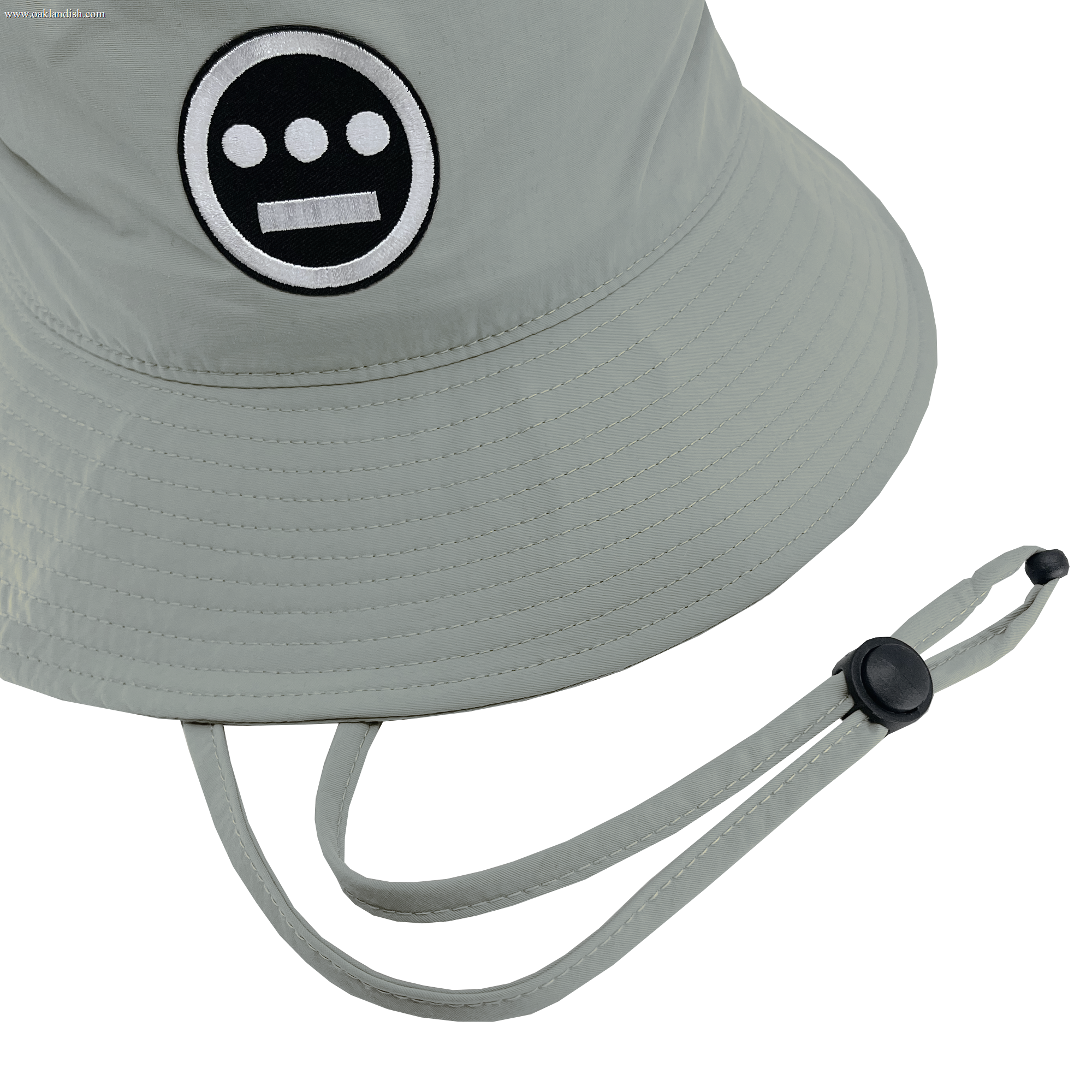 The Hieroglyphics Hiero Boonie Hat is a light gray bucket hat made from recycled nylon, showcasing a black and white embroidered logo of a circular face with two horizontal lines for eyes and one for a mouth. It features a chin strap and an adjustable black plastic toggle, merging style with sustainability as an eco-friendly accessory.