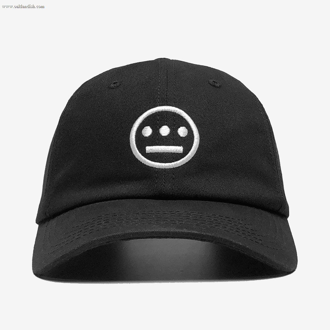 The Hiero Logo Dad Hat by Hieroglyphics is a black dad hat featuring a white, embroidered logo on the front. The logo, inspired by hieroglyphics, depicts a face with three dots for eyes and a straight line for a mouth, all enclosed in a circle. This headwear boasts a curved brim and six panels topped with a button, set against its plain white background.