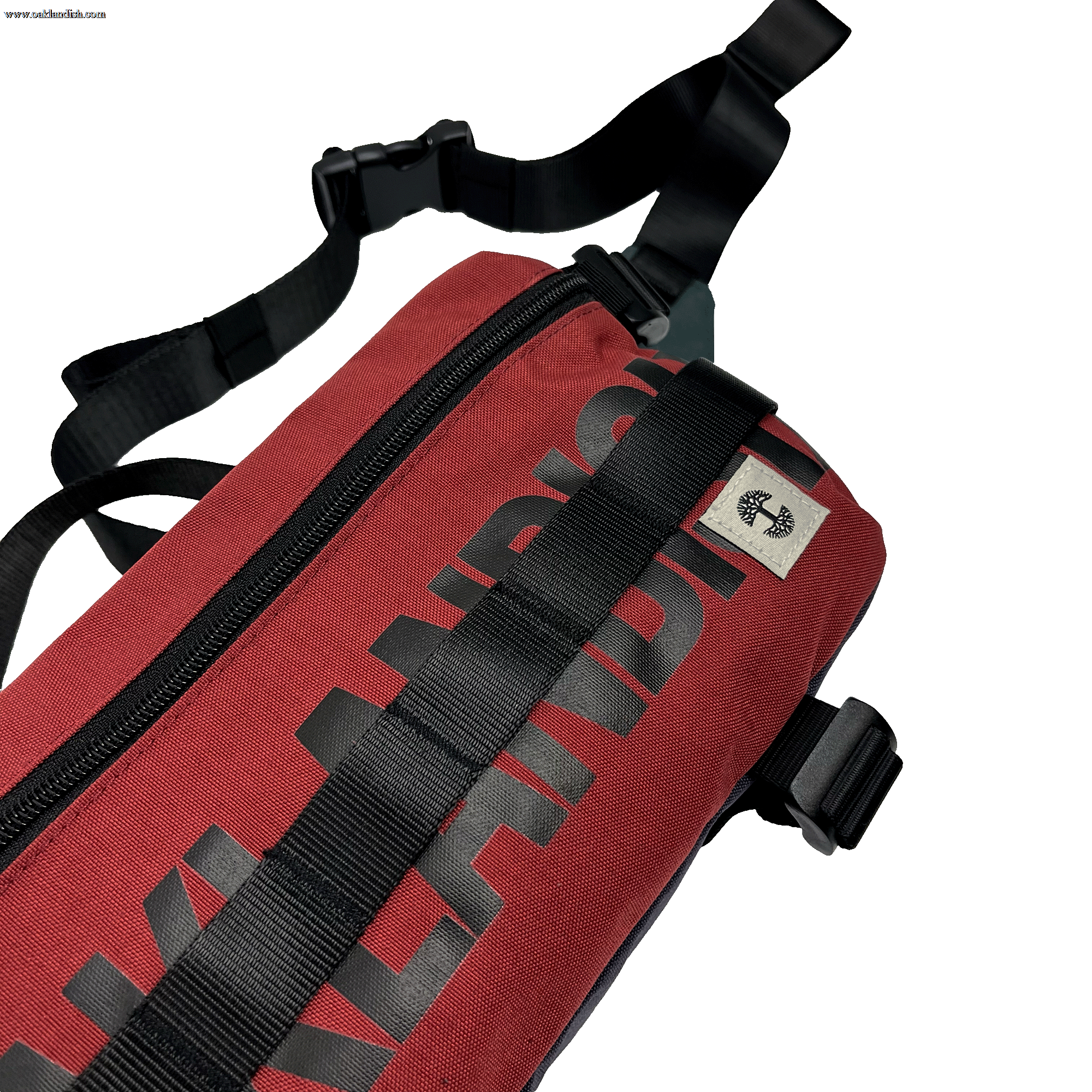 A close-up image of the Oaklandish Hip Bag, a red shoulder bag featuring black straps and a black zipper. The word "Oaklandish" is printed in bold black letters along the side of the bag. With its unisex appeal and premium details, including a small cream tag with the Oaklandish logo near the top, this versatile bag is positioned on a white background.