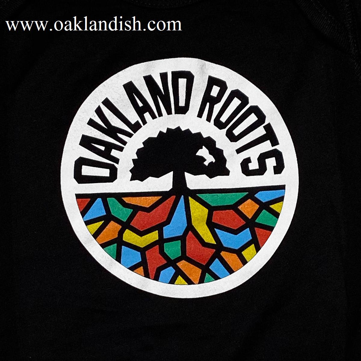 The Infant Oakland Roots SC Logo One-Piece by Oakland Roots SC features a logo design with a white circle, inside of which the words "Oakland Roots" are arched along the top. Below this text is a stylized tree design with black roots extending into a vibrant mosaic of colorful, abstract shapes in red, blue, yellow, green, and orange. Perfect for showing your support for your favorite sports club on an adorable baby romper. The background is black.