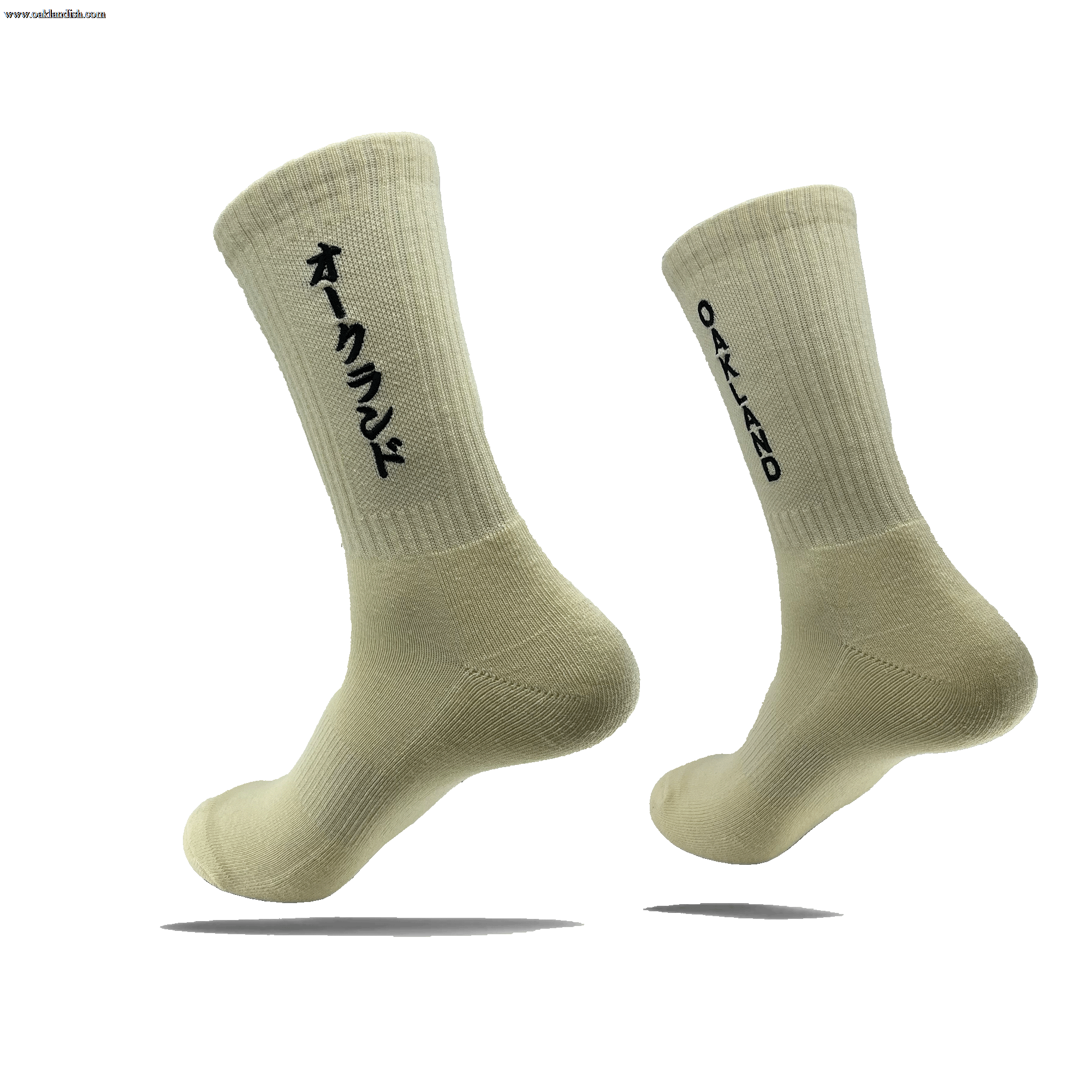 Image of the "Kanji Sock" by Oaklandish, a pair of off-white, ribbed ankle socks. The left sock displays Japanese Katakana characters in black, while the right sock features the word "OAKLAND" in black capital letters. Both cotton socks are shown against a plain white background, slightly elevated to showcase their design. Available in adult sizes.