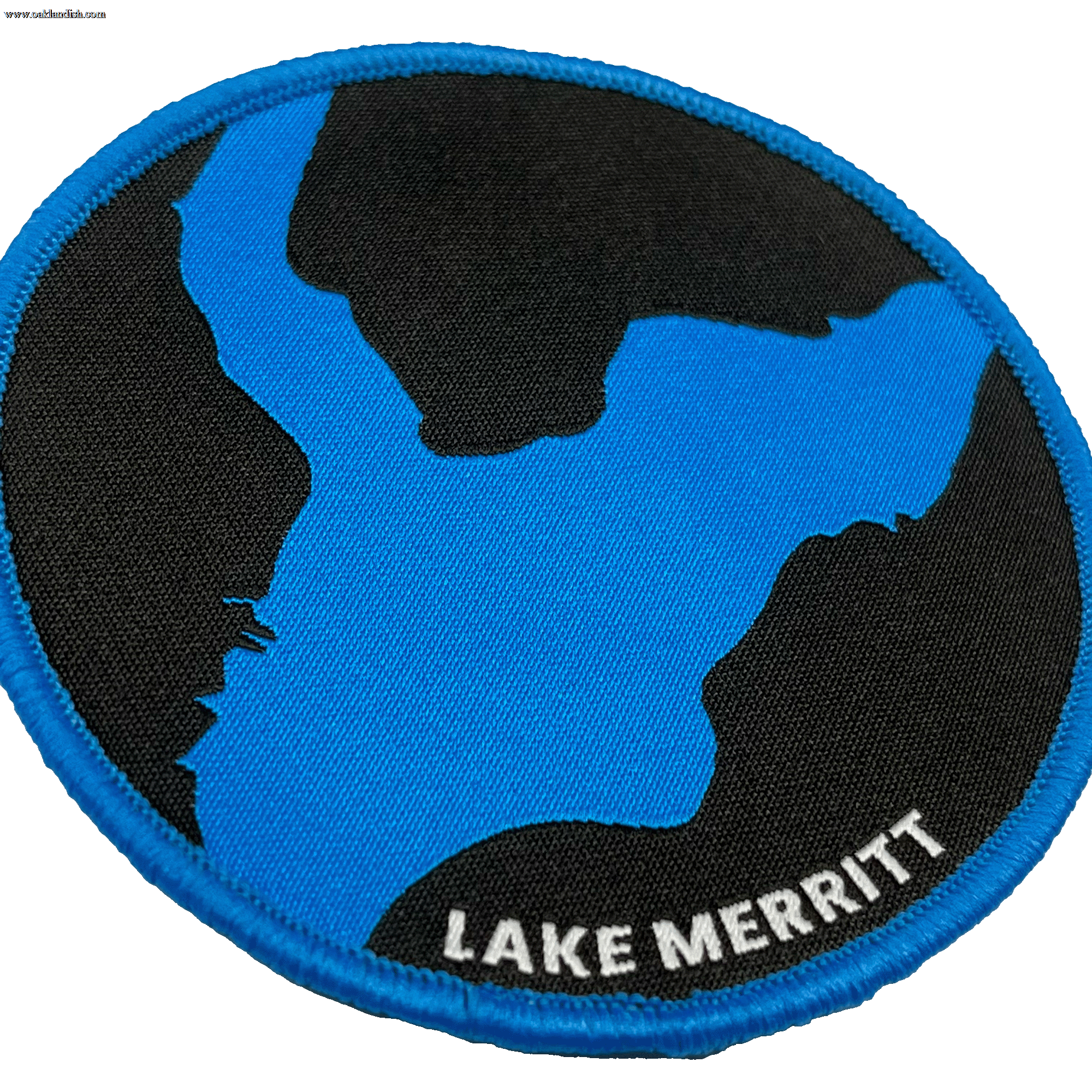 The Oaklandish Lake Merritt Patch features a rounded design with a black background showcasing a blue stylized map of Lake Merritt. The edge is trimmed in blue, and the words "LAKE MERRITT" are elegantly embroidered in white text at the bottom right of this versatile iron-on patch, making it perfect for personalizing your accessories.