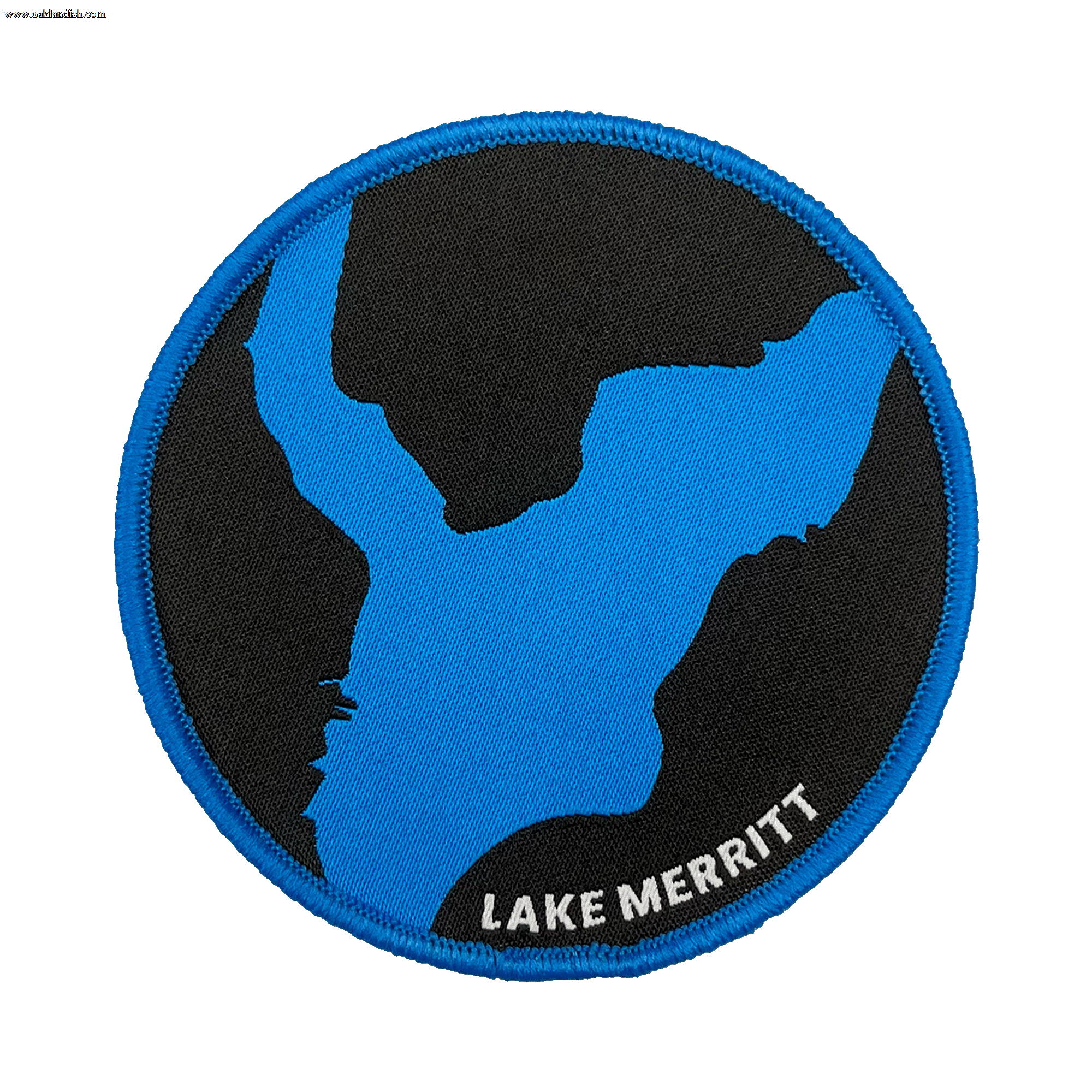 The Oaklandish Lake Merritt Patch is a circular embroidered iron-on patch that features a stylized map outline of Lake Merritt in blue against a black background, with a blue border. The label "LAKE MERRITT" is embroidered in white at the bottom right edge, making it the perfect customized patch for your accessories.